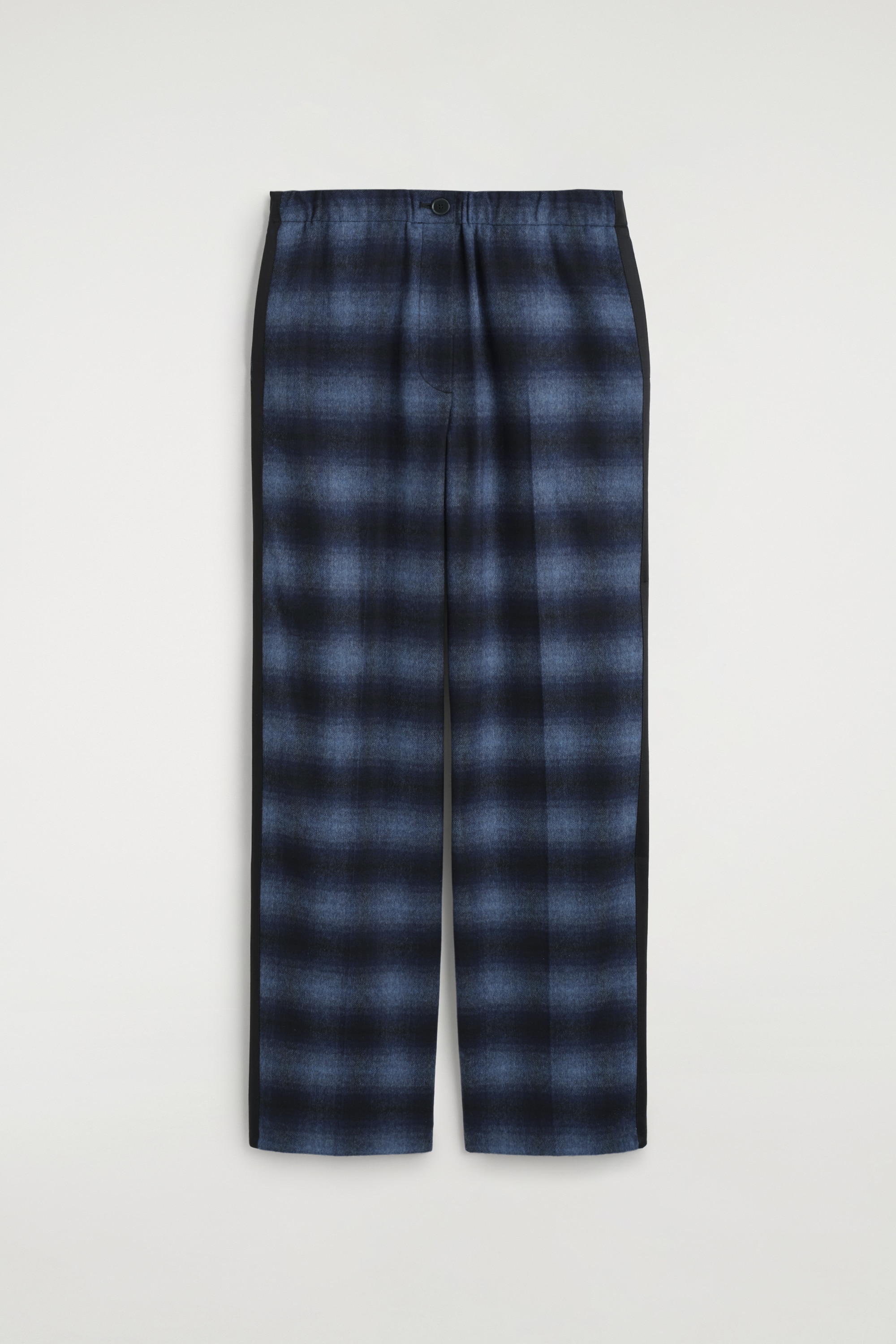 Le mannequin porte un pantalon en mÃ©lange de laine Ã  carreaux, prÃ©sentant un motif tartan bleu foncÃ© et noir, avec une bande latÃ©rale en satin noir. Le pantalon est associÃ© Ã  un haut en satin bleu marine Ã  manches longues, rentrÃ© Ã  lâintÃ©rieur, une fine ceinture en cuir noir et des chaussures en cuir noir.