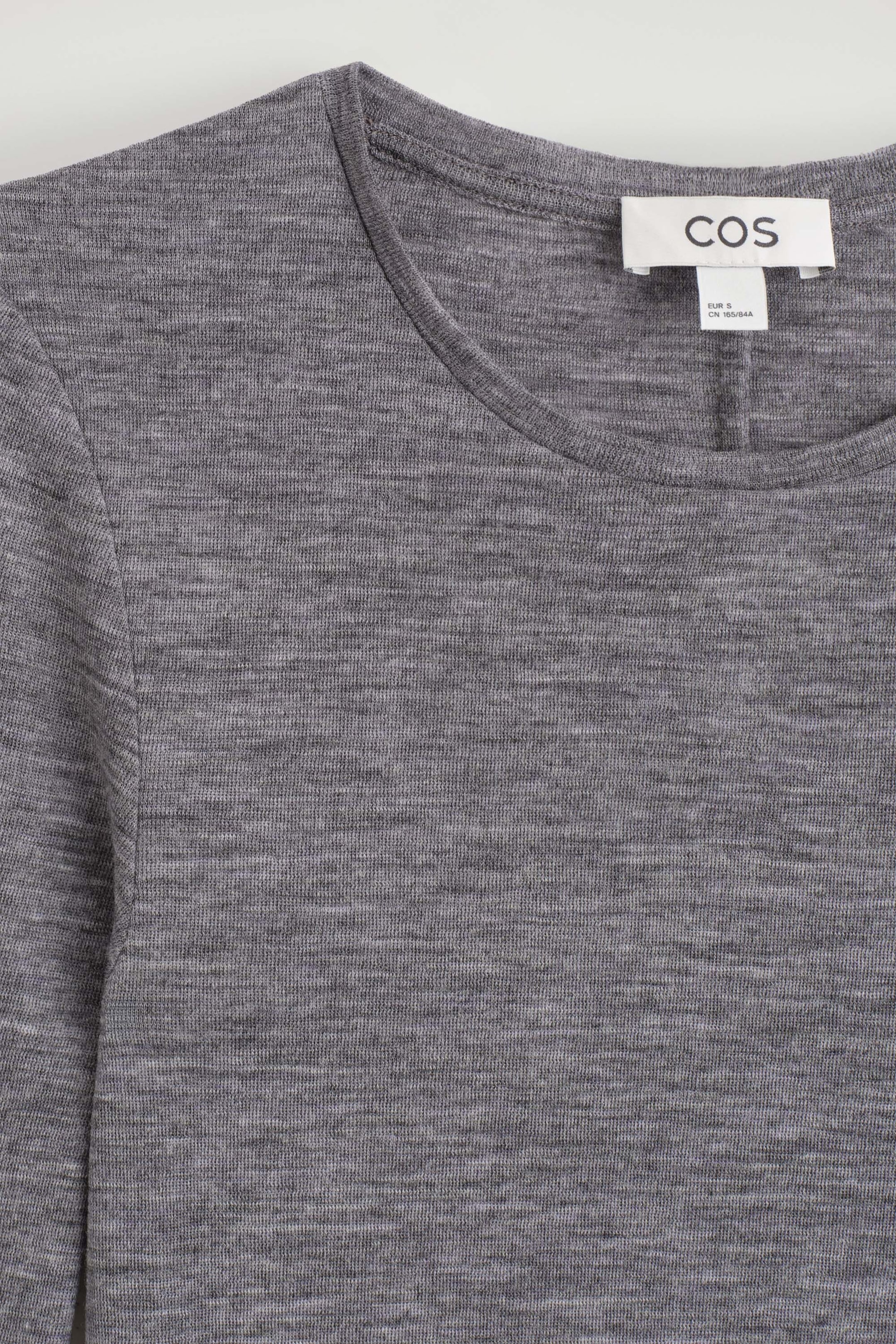 The front of this slim merino wool top shows a close-up of its heathered grey, fine-knit fabric. The top features a classic crew neck with a stitched band and a white brand label sewn inside the collar.