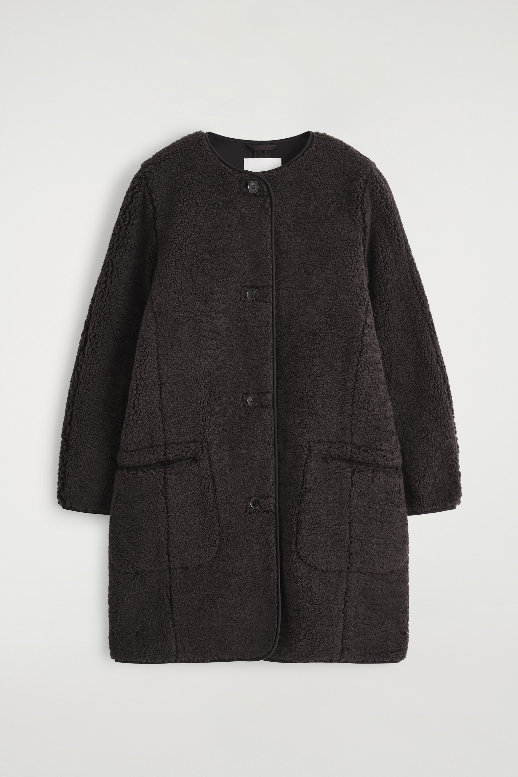 Ingrandisci l'immagine: Un cappotto marrone scuro senza colletto presenta la sua vista frontale, caratterizzato da un tessuto bouclé testurizzato. È progettato con una chiusura frontale a bottoni, due tasche applicate e un piping scuro liscio lungo tutti i bordi.