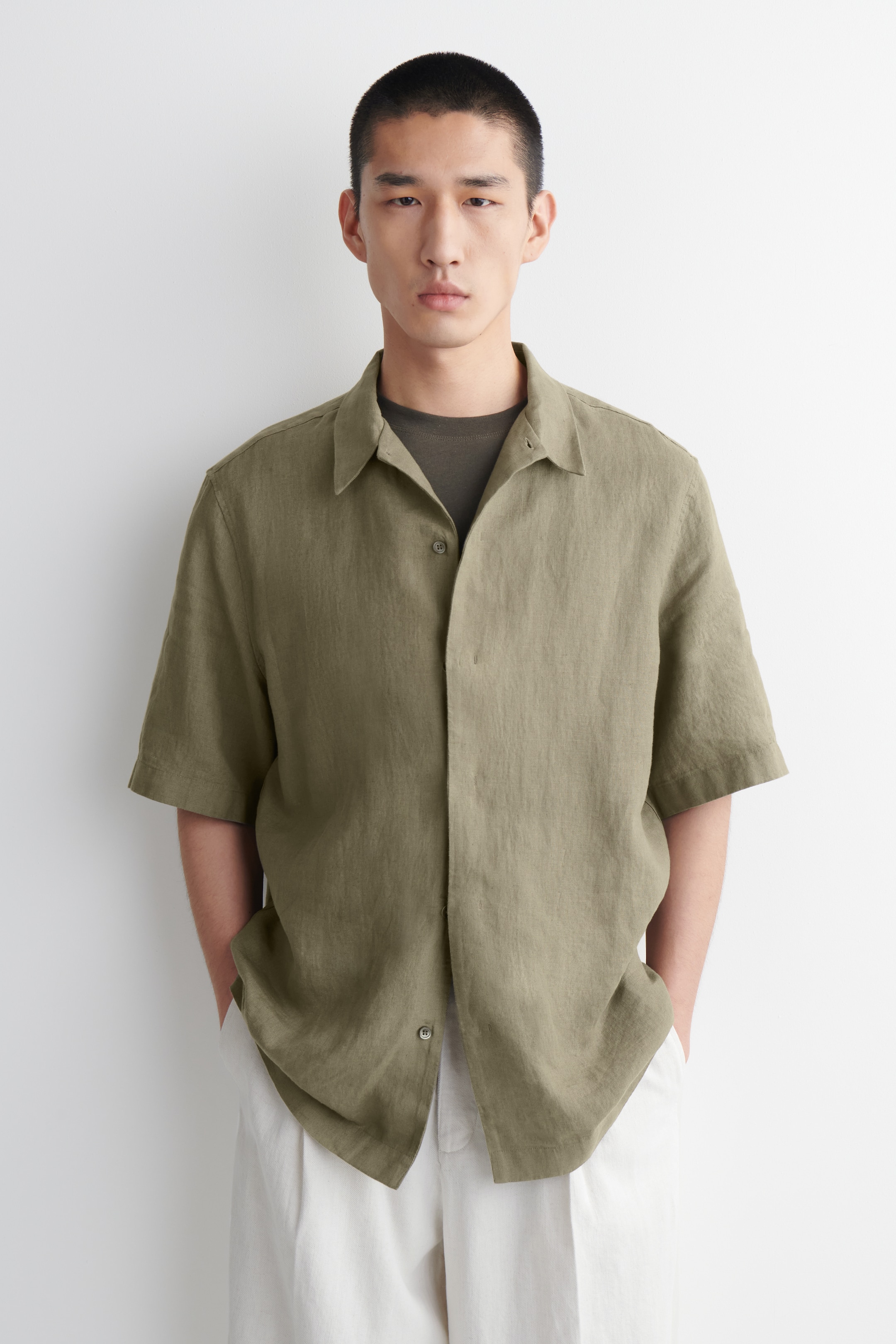 Ver imagen más grande: Se lleva puesta una camisa de manga corta de color verde oliva, orientada hacia adelante, sobre una camiseta verde oscuro. Presenta un cuello extendido y mangas cortas. La camisa está parcialmente desabotonada, mostrando un ajuste relajado y un tejido texturizado.