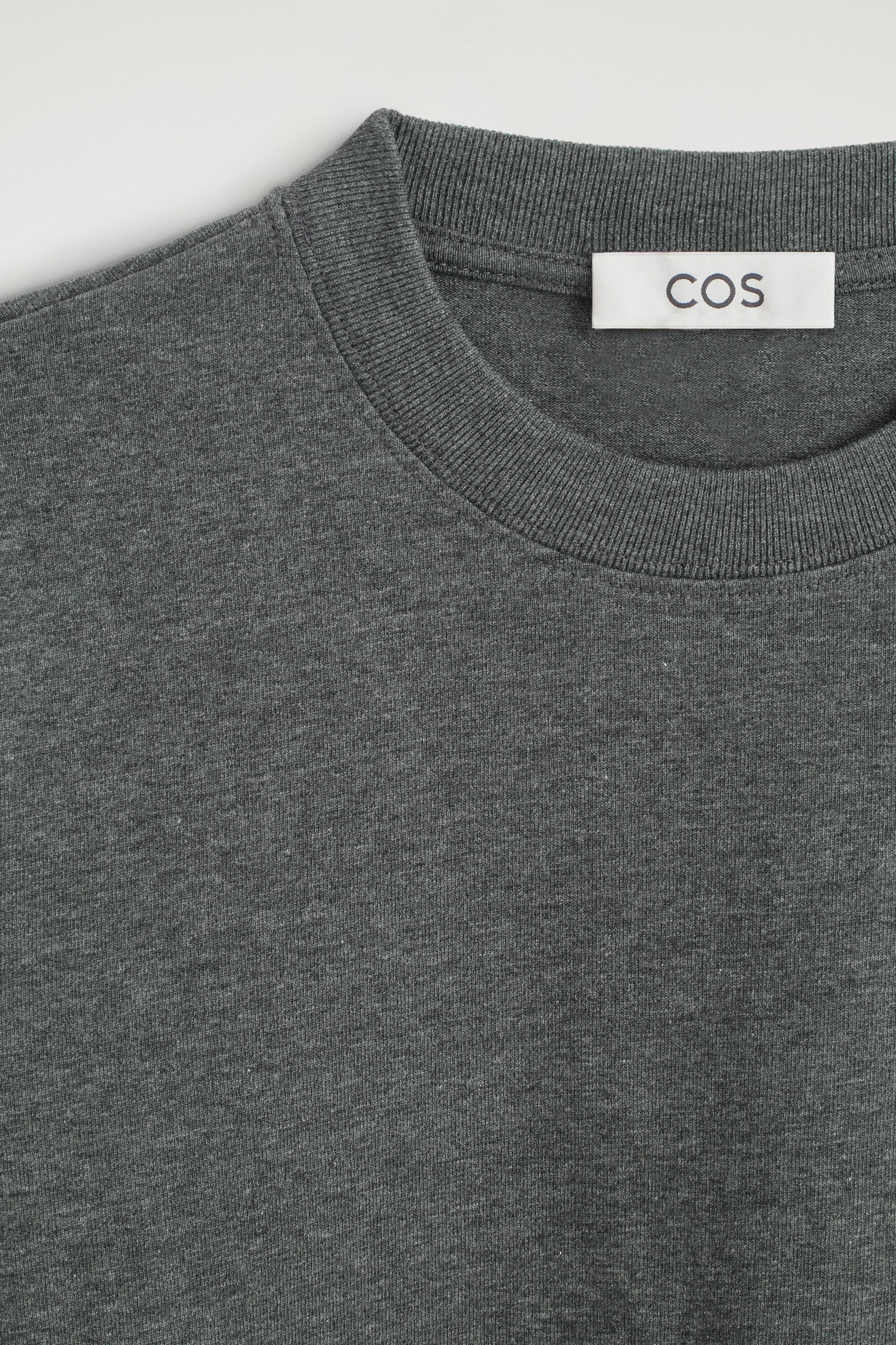 The clean-cut long-sleeved T-shirt is shown in a close-up front view, highlighting its dark gray melange fabric and ribbed crewneck. A white brand label reading "COS" is visible stitched inside the back collar.