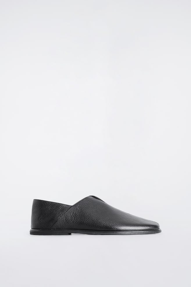 Ein schwarzer Leder-Mule ruht in seitlicher Profilansicht auf seiner flachen Sohle und zeigt sein strukturiertes genarbtes Leder, die mandelförmige Spitze und den V-förmigen Spann. Das Slip-on-Design verfügt seitlich über ein überlappendes Lederpanel. - 1