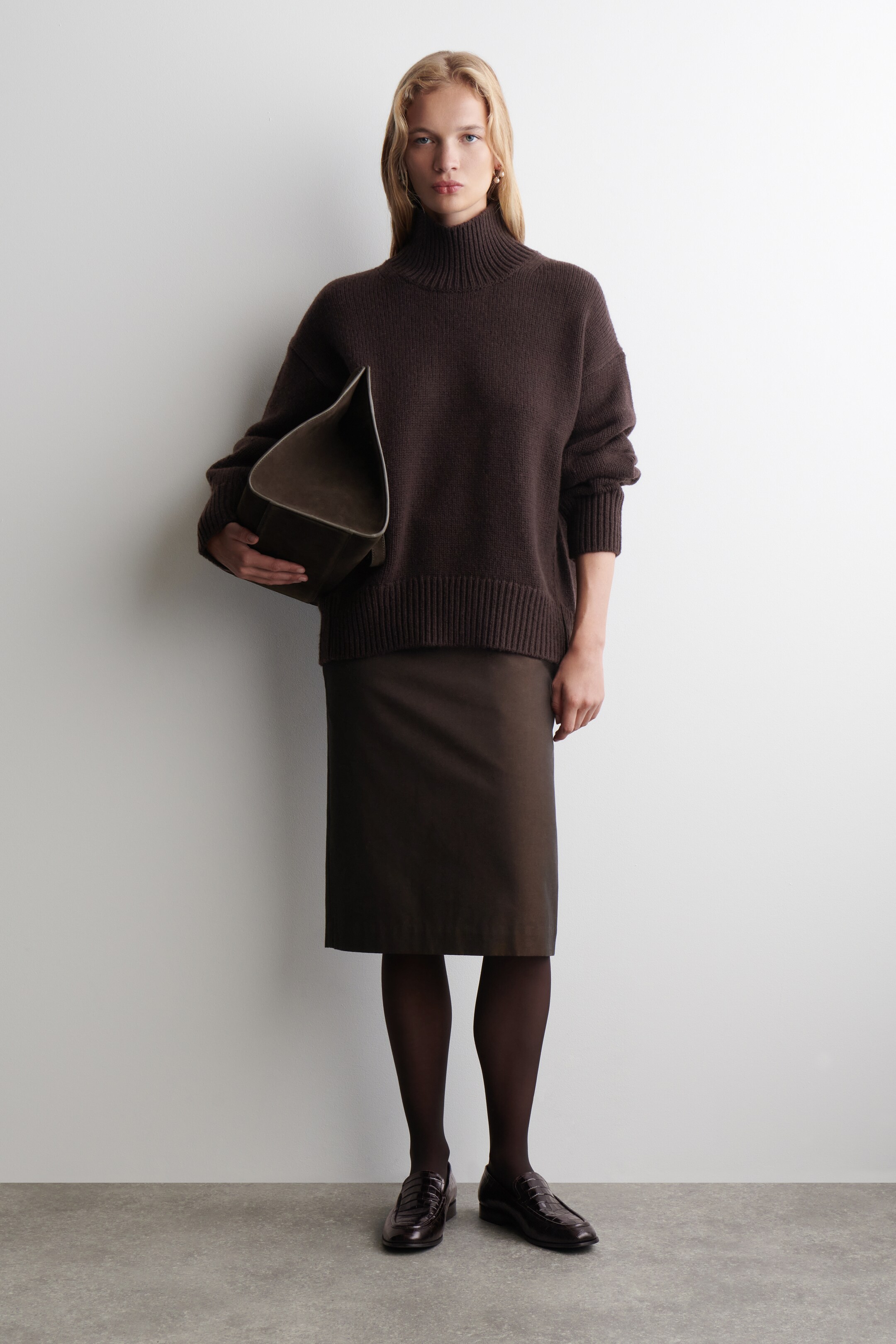 View larger image: A dark brown turtleneck sweater, featuring a relaxed fit, ribbed collar, cuffs, and hem, is being worn with a dark brown midi-length pencil skirt. A structured dark brown bag is being held against the body, and opaque dark tights with dark brown loafers are completing the outfit.