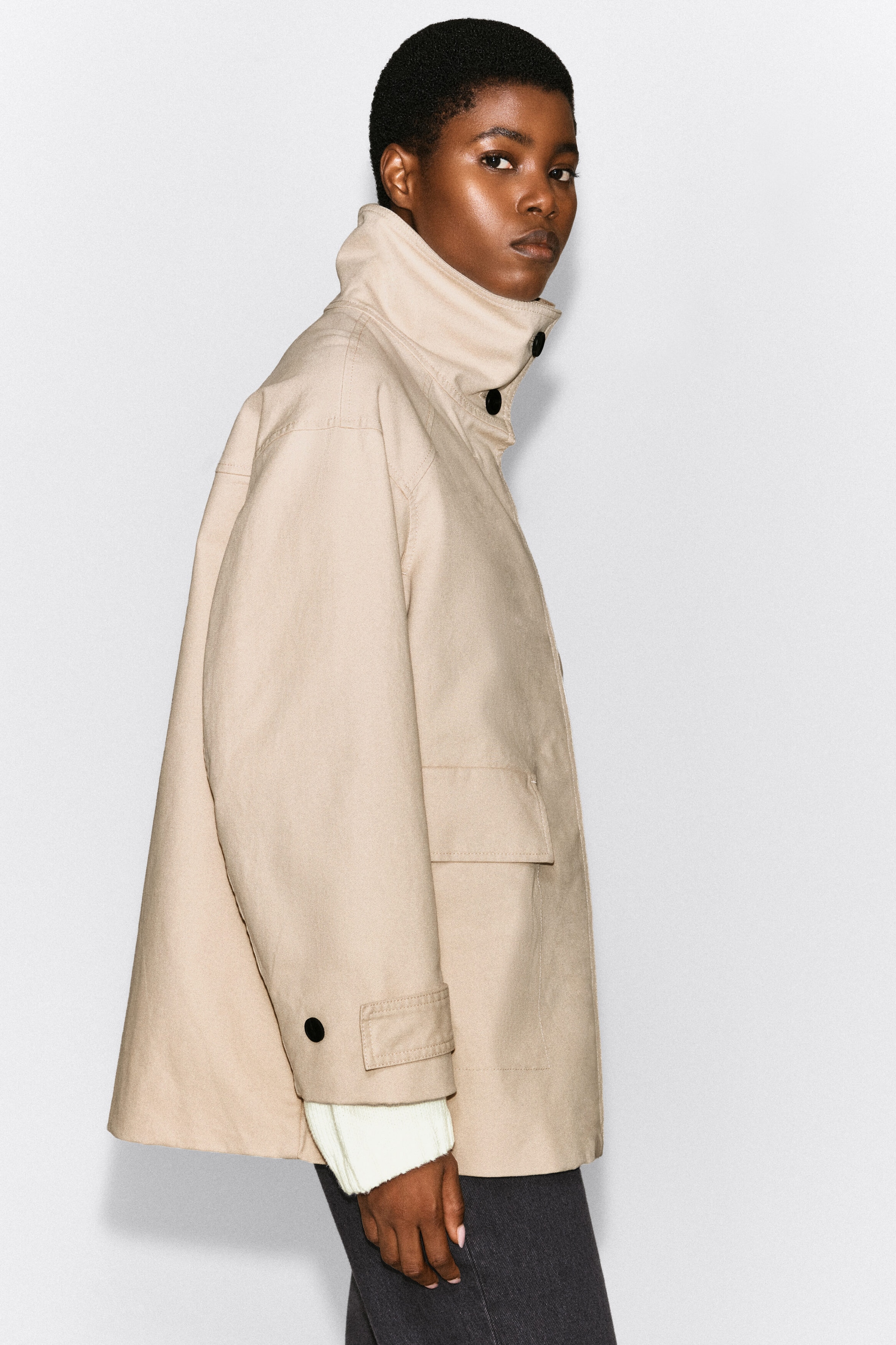 The model is wearing a beige cotton twill funnel-neck jacket featuring a high buttoned collar, a side flap pocket, and button-tab cuffs. A cream-coloured ribbed knit sleeve peeks out from the cuff, layered beneath the jacket. The model is wearing a beige cotton twill funnel-neck jacket featuring a high buttoned collar, a side flap pocket, and button-tab cuffs. A cream-coloured ribbed knit sleeve peeks out from the cuff, layered beneath the jacket.