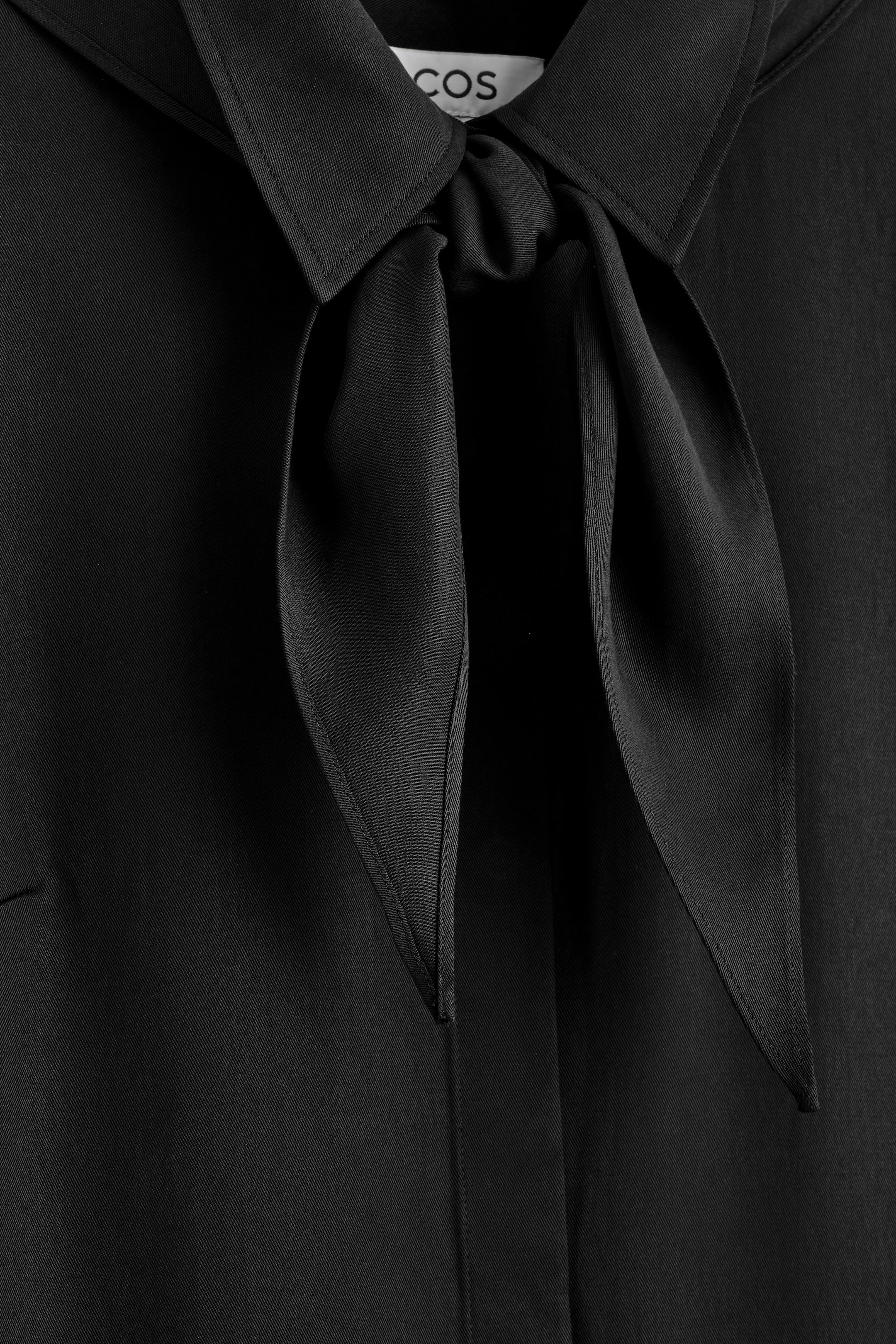 Visualizza immagine più grande: Un indumento nero presenta un colletto a punta, con due strisce di tessuto annodate sul davanti, creando un dettaglio a cravatta. Il tessuto mostra una sottile trama a intreccio diagonale.