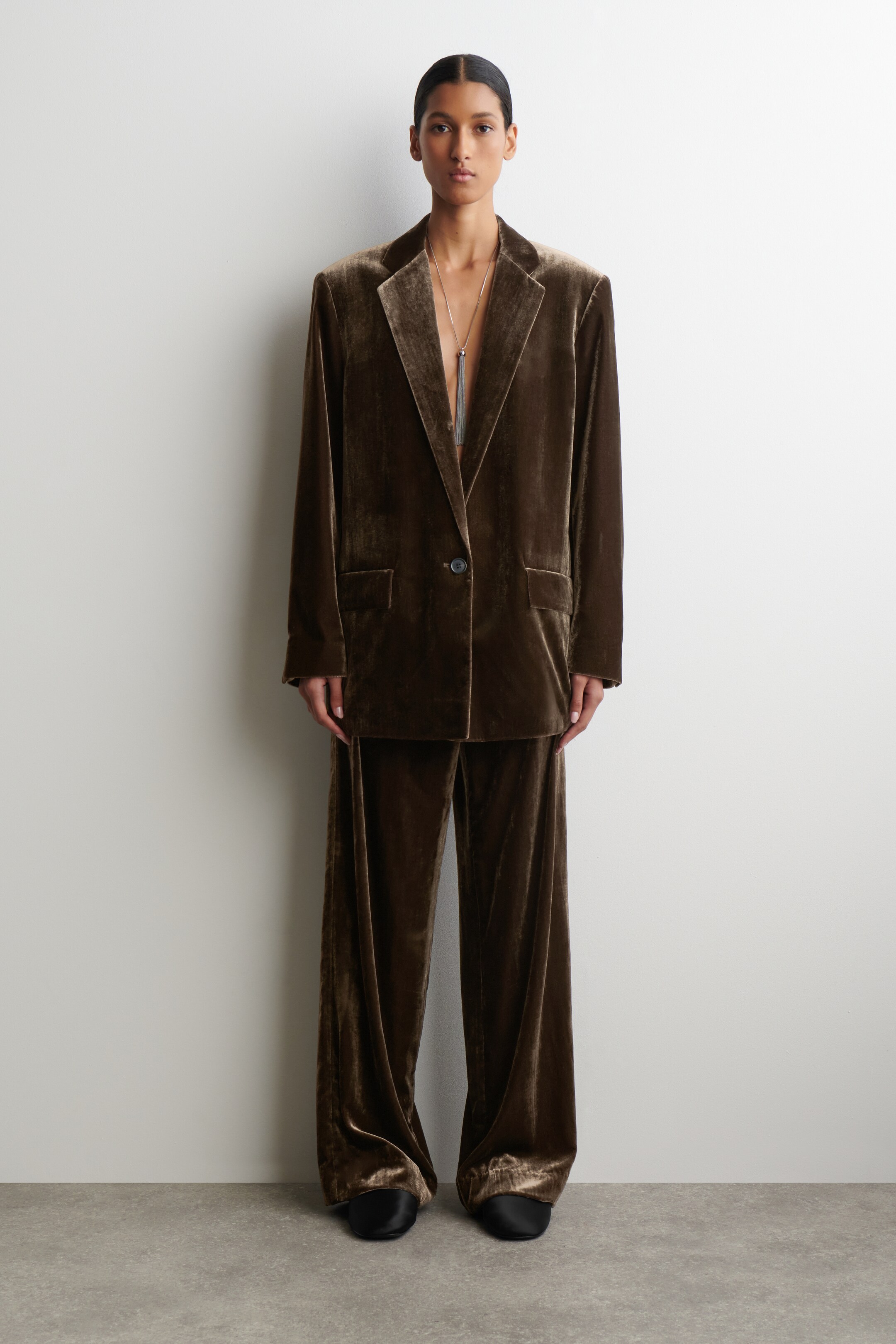 Agrandir l'image: Un costume en velours marron, présenté de face, est doté d'une veste surdimensionnée avec des revers à encoches, une fermeture à un bouton et deux poches à rabat, assorti d'un pantalon à jambes larges. Un long collier à pampilles en argent se drape sur la veste ouverte, et des chaussures noires sont visibles sous le pantalon.