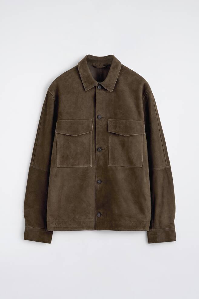 Eine dunkelbraune Wildlederjacke zeigt ihre Vorderansicht, mit einem spitzen Kragen und einer Knopfleiste mit dunklen, runden Knöpfen. Zwei große aufgesetzte Taschen mit Klappen sind auf der Brust sichtbar, und lange Ärmel mit Einknopfmanschetten vervollständigen das Design. - 1