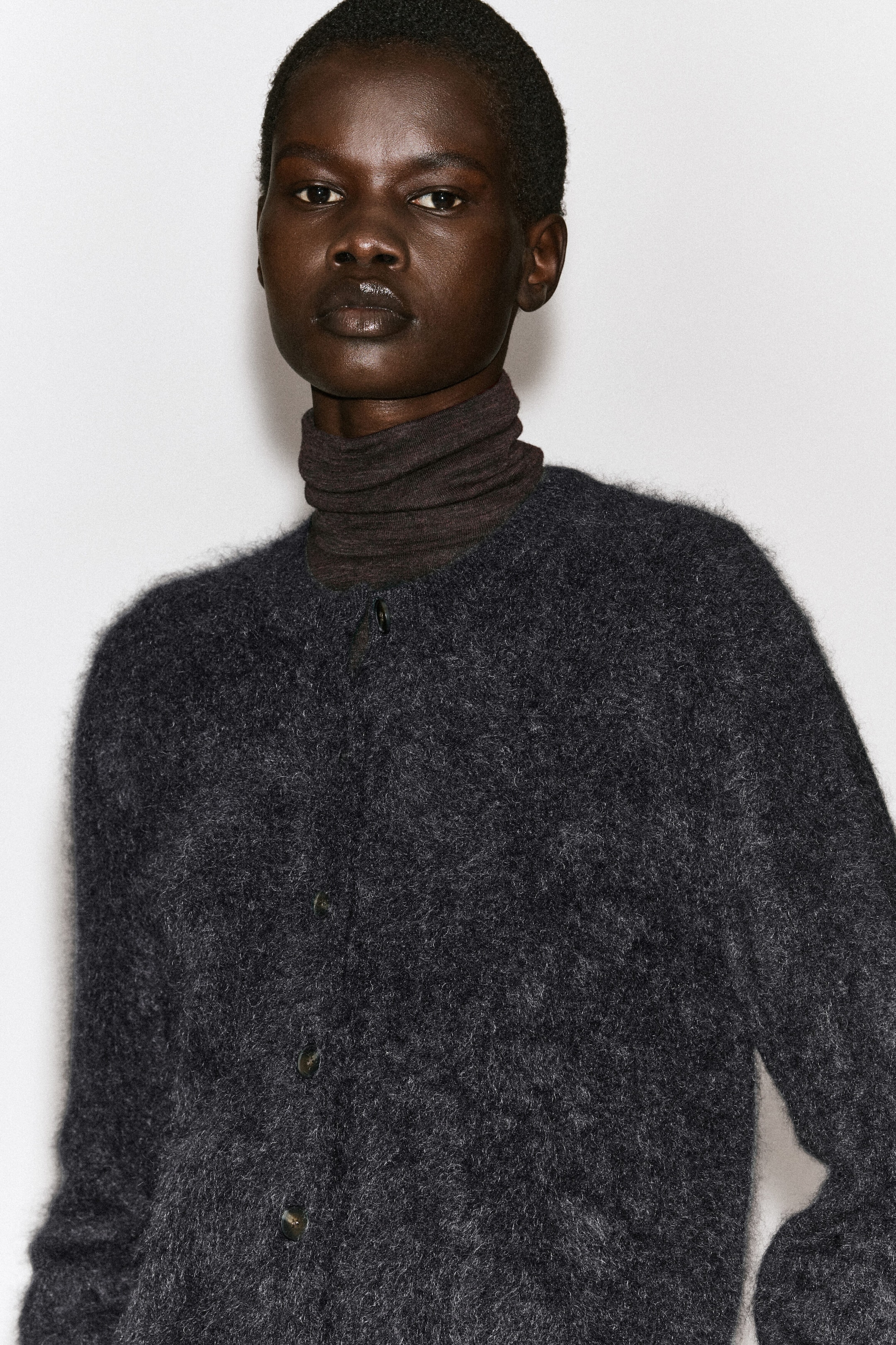 The model is wearing a dark grey brushed pure cashmere crew-neck cardigan layered over a dark brown turtleneck top. The cardigan features a fuzzy, textured finish and a button-front closure. The model is wearing a dark grey brushed pure cashmere crew-neck cardigan layered over a dark brown turtleneck top. The cardigan features a fuzzy, textured finish and a button-front closure.