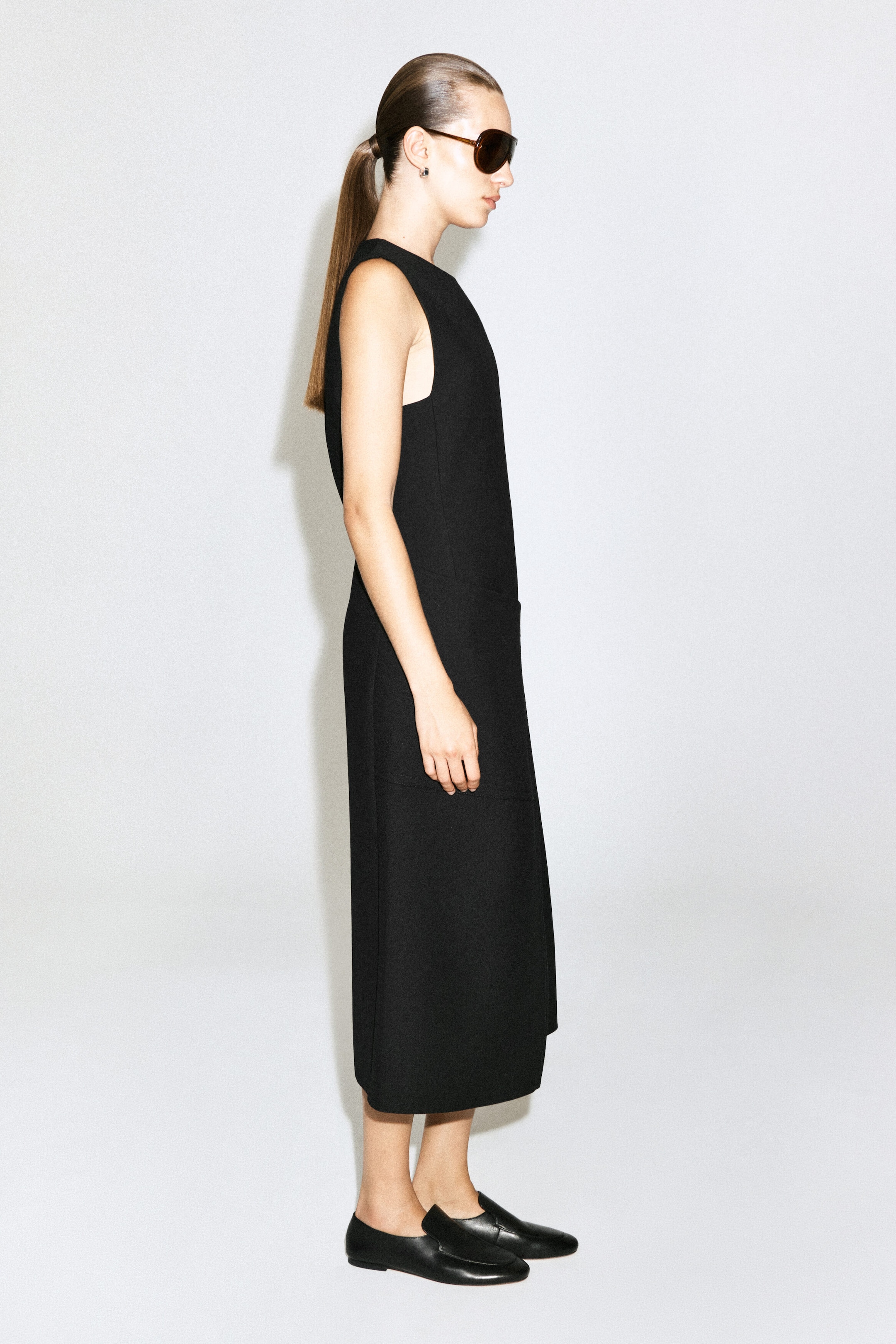 A black, sleeveless, double-faced wool midi dress is being worn by the model, who is standing in profile. The structured, straight-cut dress features a high neckline and is styled with black leather loafers and sunglasses.