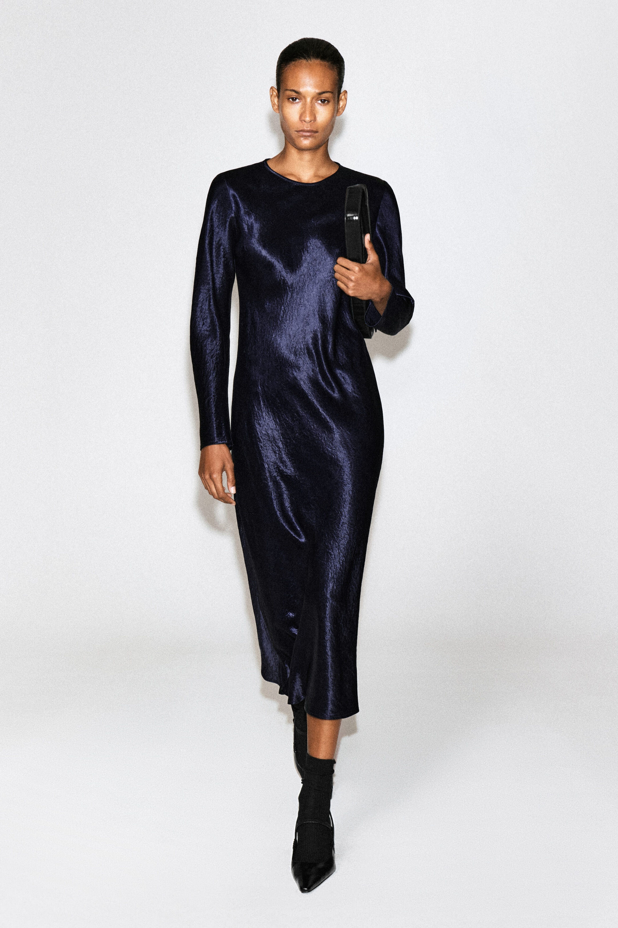 A dark navy blue satin, long-sleeved maxi dress with a textured metallic sheen is worn by the model. The dress features a round neckline and is styled with a black clutch bag held under the model's arm and black pointed-toe shoes. A dark navy blue satin, long-sleeved maxi dress with a textured metallic sheen is worn by the model. The dress features a round neckline and is styled with a black clutch bag held under the model's arm and black pointed-toe shoes.