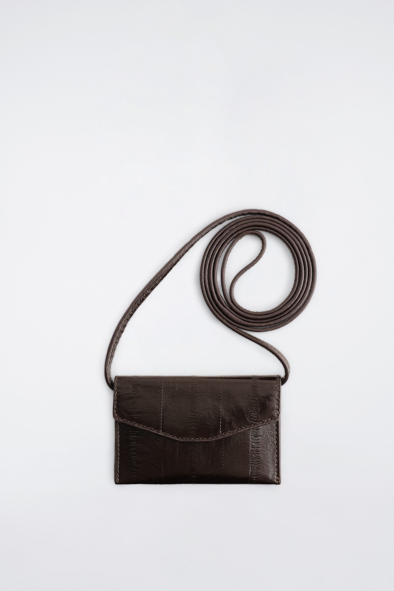 An eel-effect leather card holder is shown facing forward, featuring a glossy dark brown surface with vertical textured panels and a curved front flap. A long, thin matching leather strap extends from the top corners and is neatly coiled above the card holder.