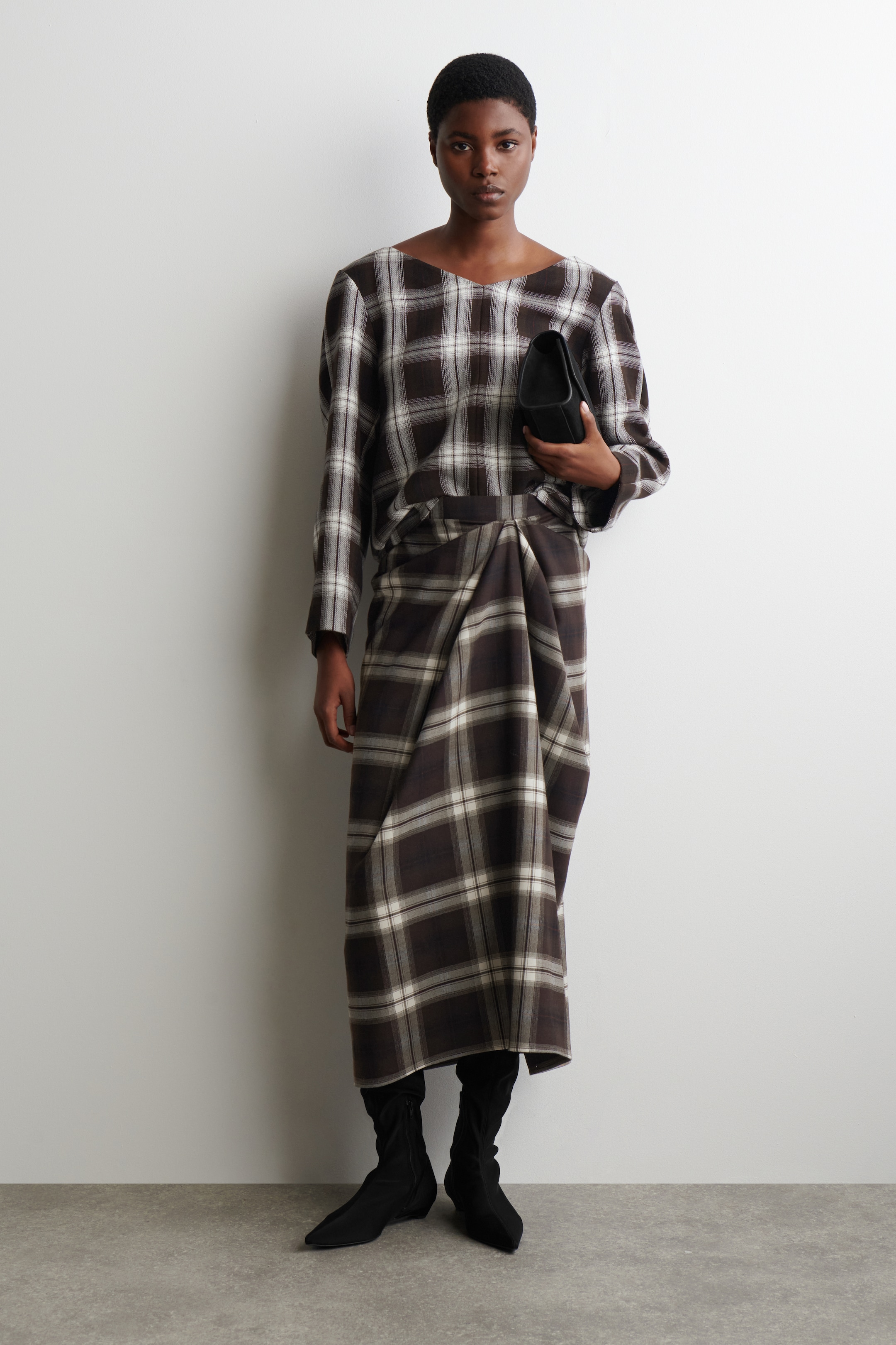 Agrandir l'image: Un haut à carreaux marron et blanc, avec une large encolure et des manches longues, est porté par le mannequin. Elle porte également une jupe midi assortie à carreaux avec des détails drapés et tient une pochette noire.