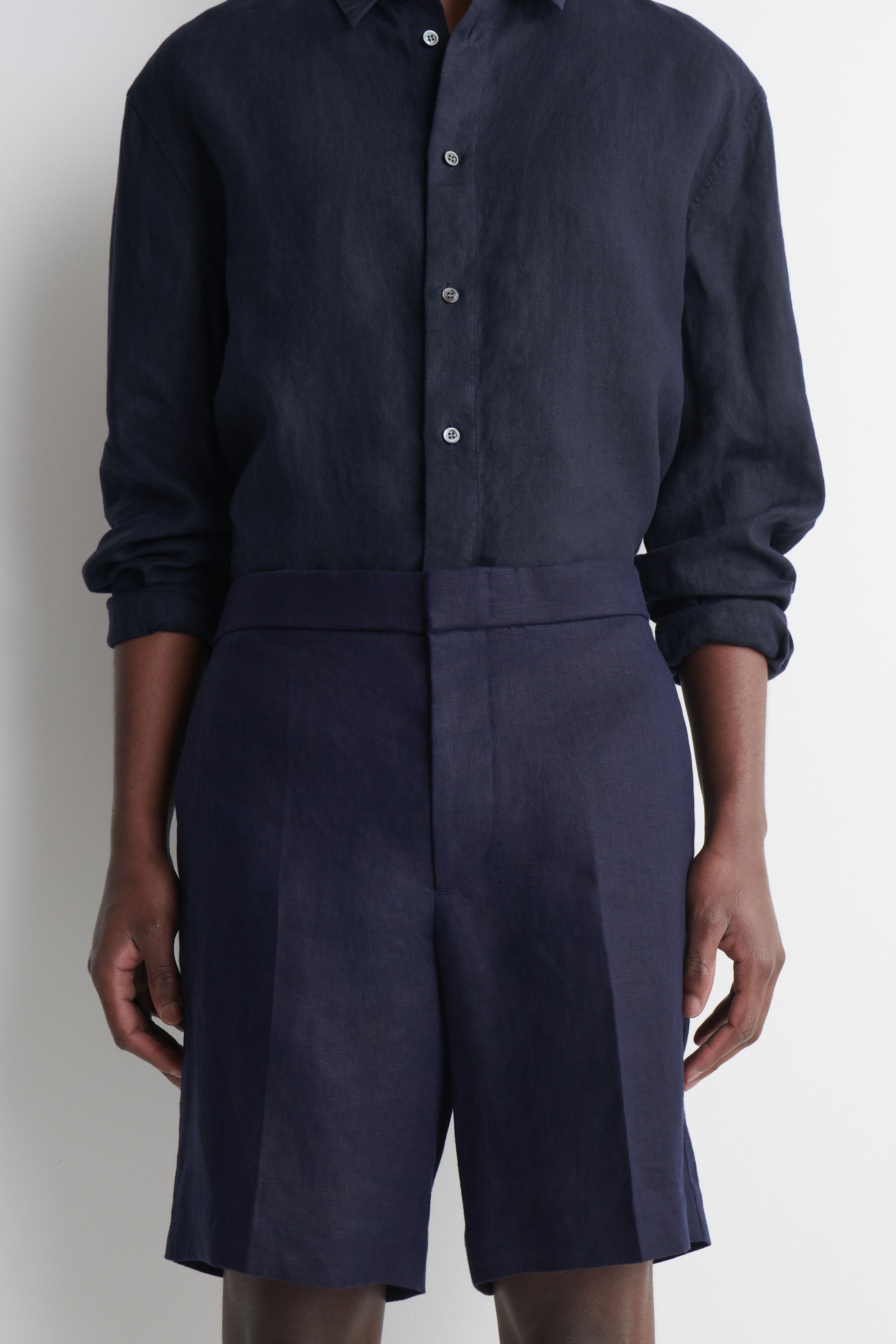 Visualizza immagine più grande: Una camicia di lino blu navy a vestibilità rilassata è indossata, caratterizzata da bottoni chiari e maniche lunghe arrotolate, ed è infilata in shorts di lino blu navy a vita alta con una piega frontale visibile.
