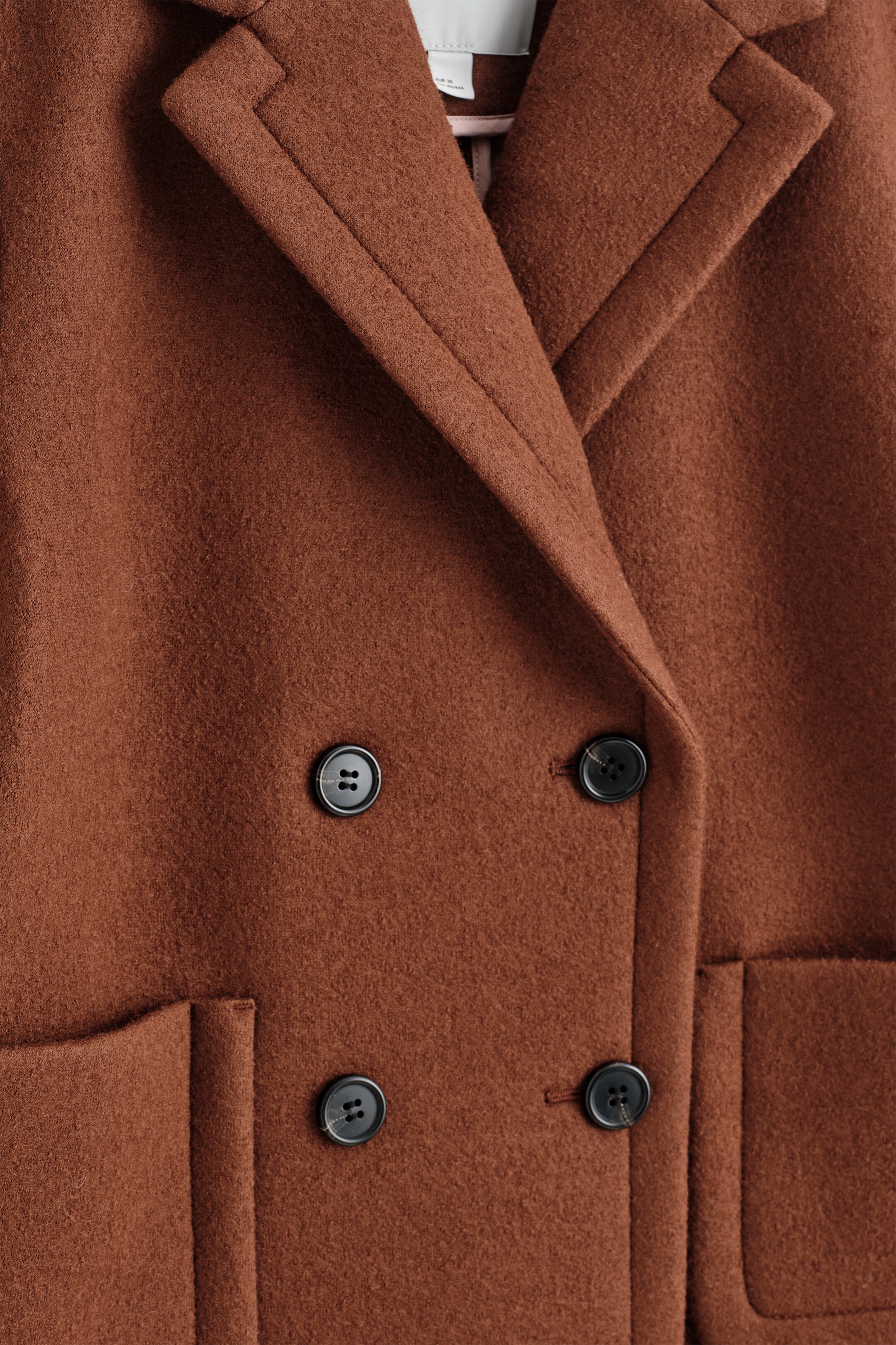 View larger image: A brown felted coat is featuring notch lapels, a double-breasted front closure with four black four-hole buttons, and two visible patch pockets.