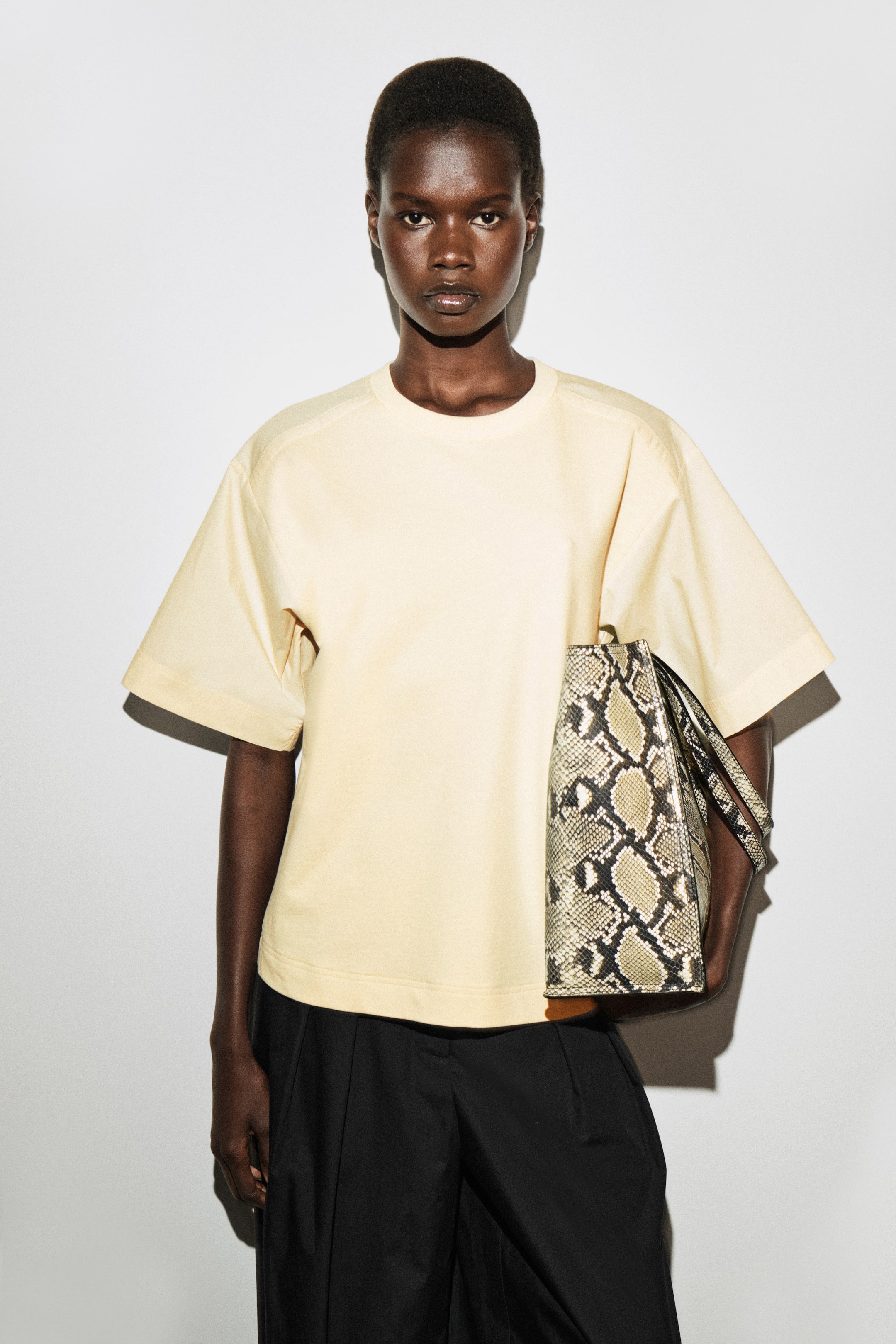 Größeres Bild ansehen: Ein hellgelbes T-Shirt mit Rundhalsausschnitt präsentiert sich in einer lockeren Passform mit überschnittenen Schultern und weiten, kurzen Ärmeln. Eine Tragetasche mit Schlangenhautmuster, die zwei obere Griffe aufweist, wird an der Seite gehalten.