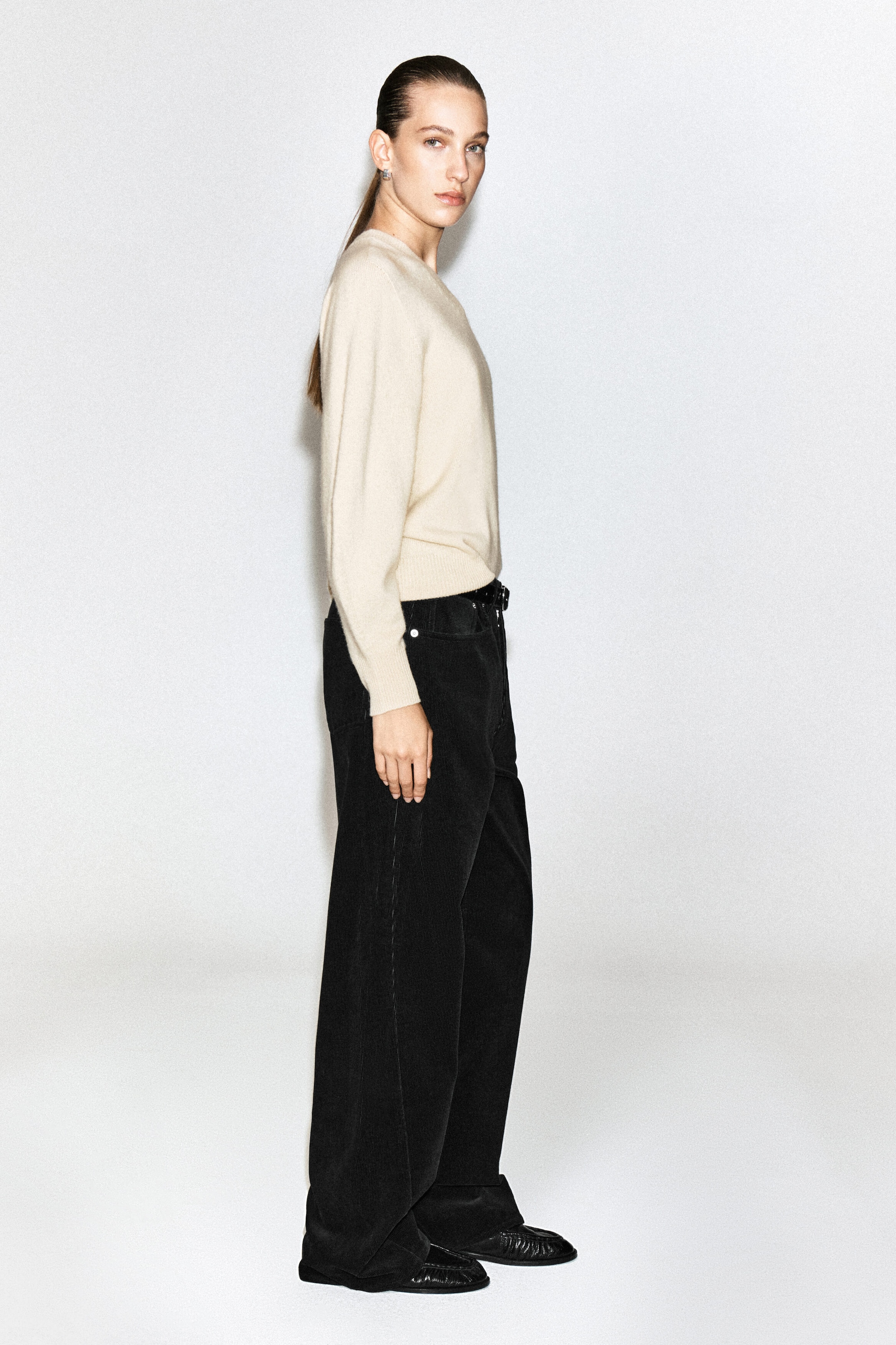The model is wearing black facade corduroy straight-leg jeans, showcasing their textured ribbed fabric, relaxed fit and high-rise waist. The jeans are styled with a tucked-in cream long-sleeved sweater and black leather shoes. The model is wearing black facade corduroy straight-leg jeans, showcasing their textured ribbed fabric, relaxed fit and high-rise waist. The jeans are styled with a tucked-in cream long-sleeved sweater and black leather shoes.