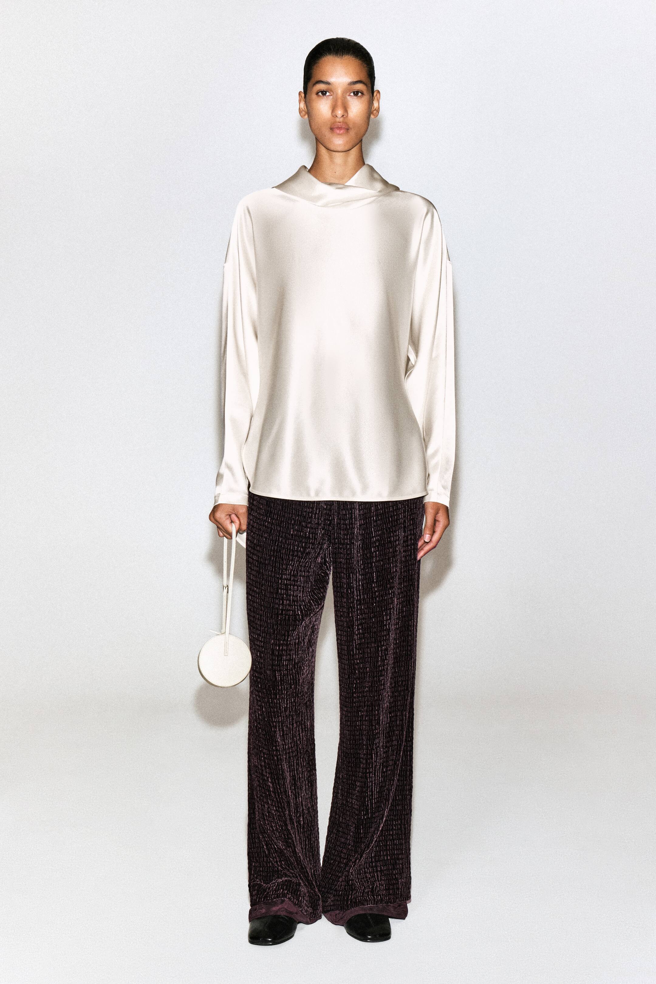 View larger image: A cream-coloured satin blouse featuring a draped cowl neck and long sleeves is shown from the front, draping loosely over the torso. It is styled with dark purple, textured wide-leg trousers and a small, circular off-white wristlet bag.