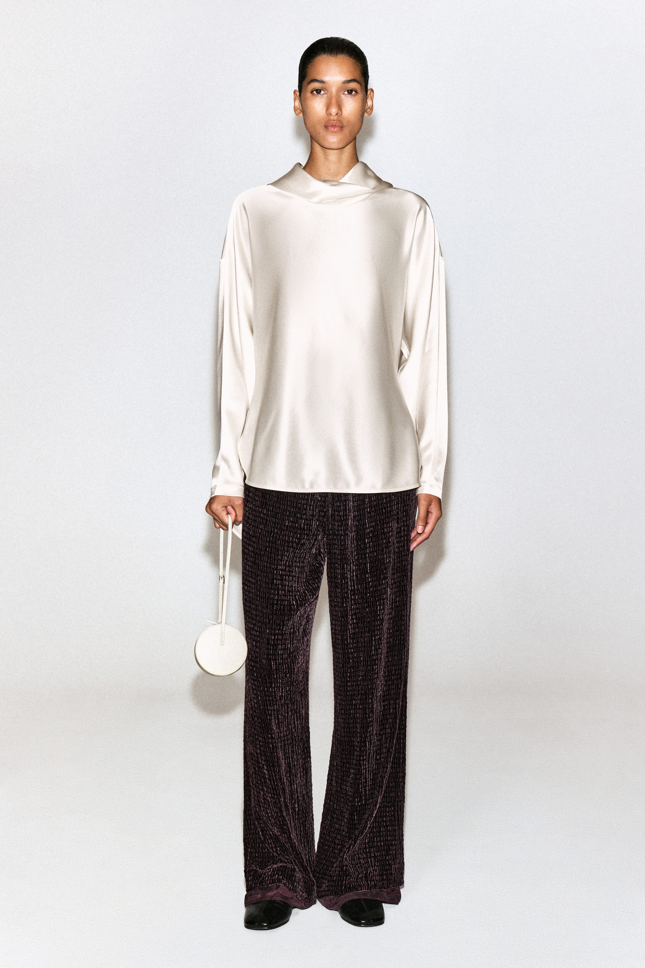 A cream-colored satin blouse featuring a draped cowl neck and long sleeves is shown from the front, hanging loosely over the torso. It is styled with dark purple textured wide-leg trousers and a small circular off-white wristlet bag. A cream-colored satin blouse featuring a draped cowl neck and long sleeves is shown from the front, hanging loosely over the torso. It is styled with dark purple textured wide-leg trousers and a small circular off-white wristlet bag.