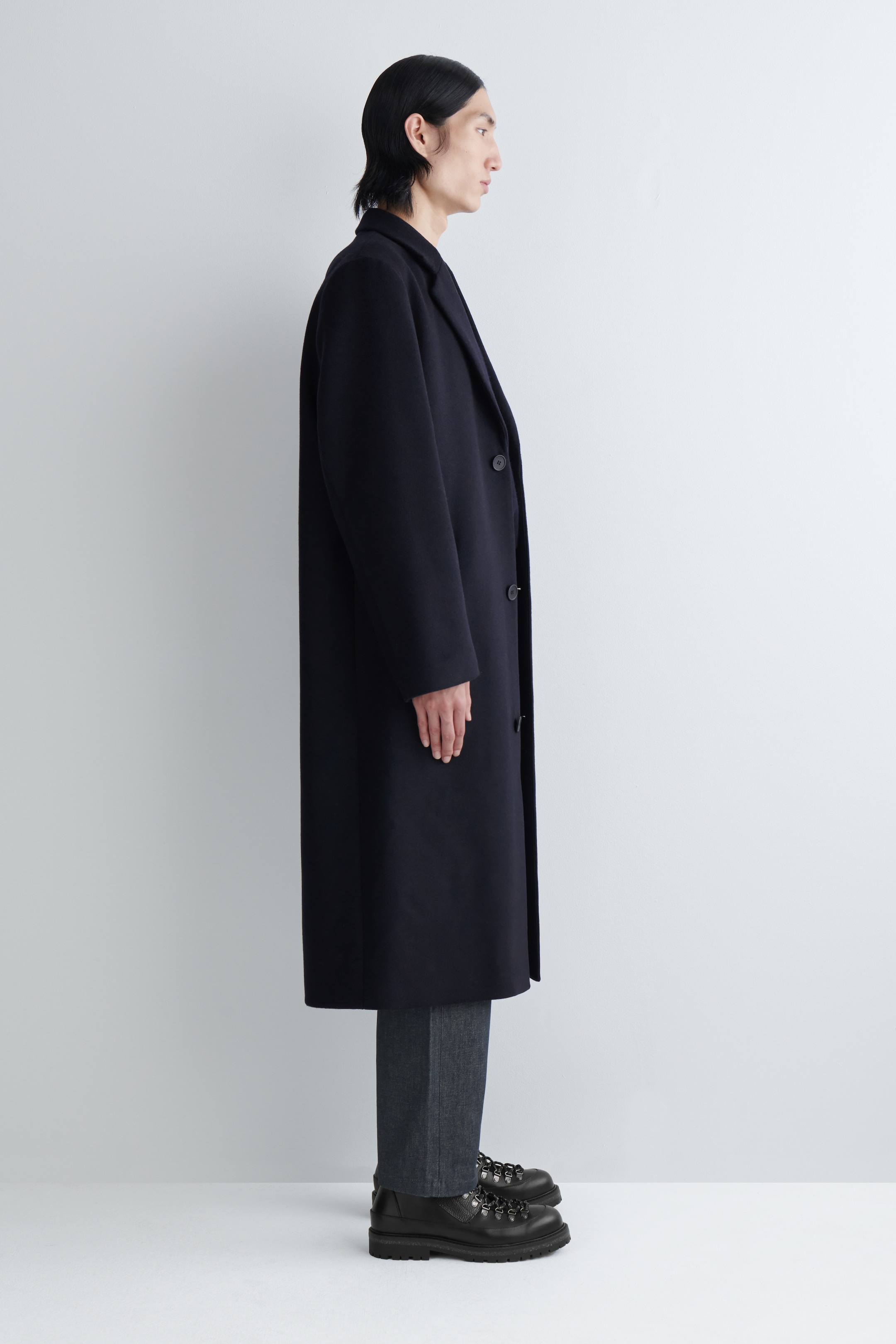 Agrandir l'image: Un manteau long de couleur sombre est présenté en vue de profil, avec des revers crantés et une fermeture à trois boutons simple boutonnage. Le manteau présente une coupe droite et décontractée et s'étend en dessous des genoux.