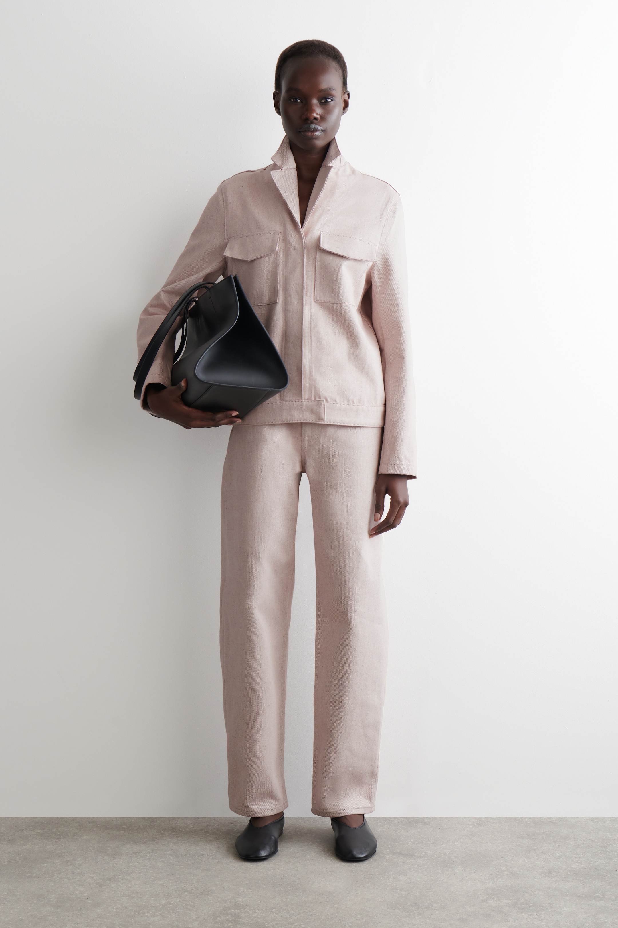 Ver imagem maior: Um conjunto rosa claro a condizer, usado de frente, apresenta um casaco com decote em V e dois bolsos de aba no peito, e calças de perna reta. Uma mala preta sem estrutura está a ser segurada, completando o visual com sapatos rasos pretos.