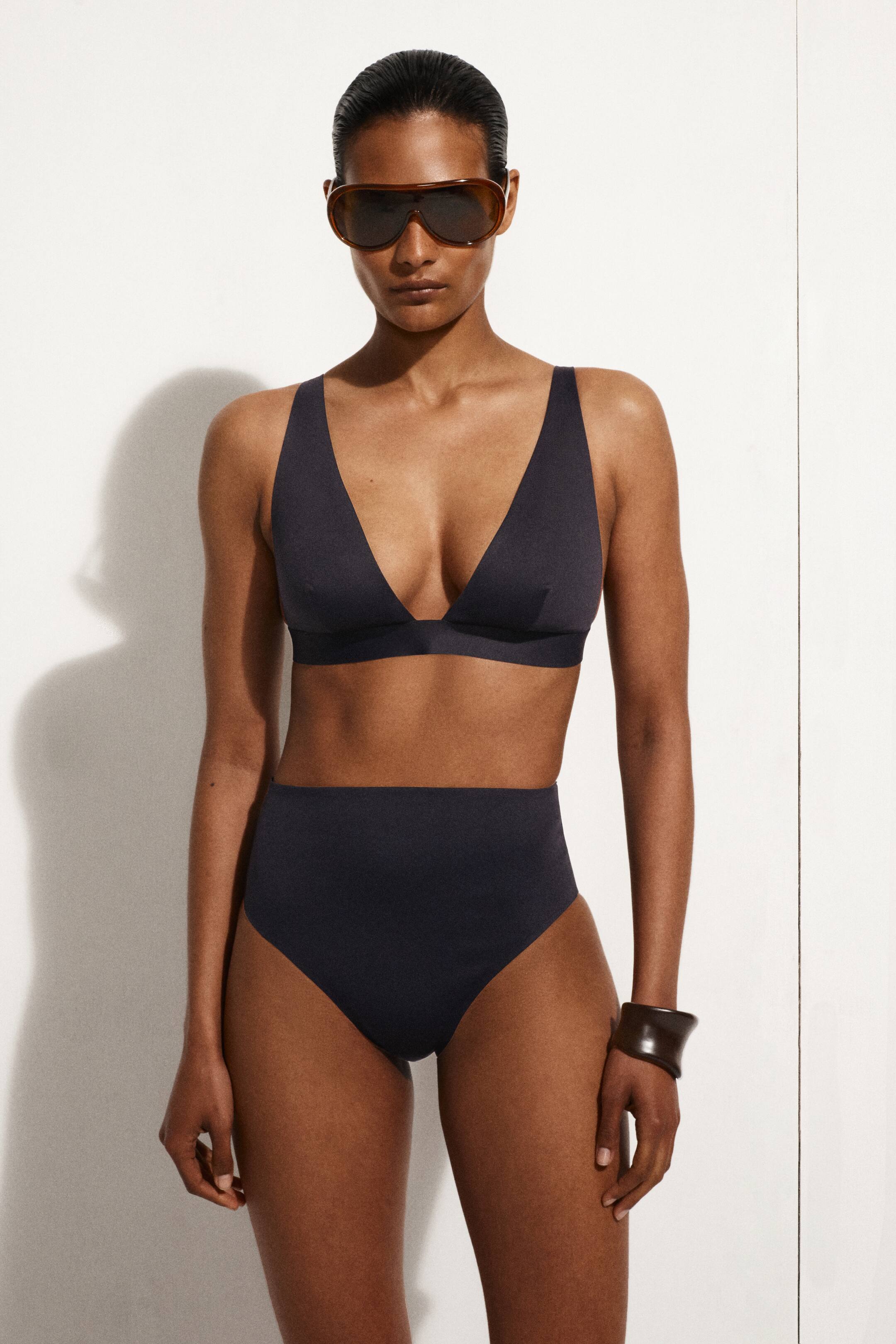 Visa större bild: Ett mörkgrått tvådelat baddräkt presenteras, med en bralette-stil topp med triangulära kupor och en djup V-ringning, samt ett brett band under bysten. Botten är en högmidjad trosa som erbjuder full täckning.