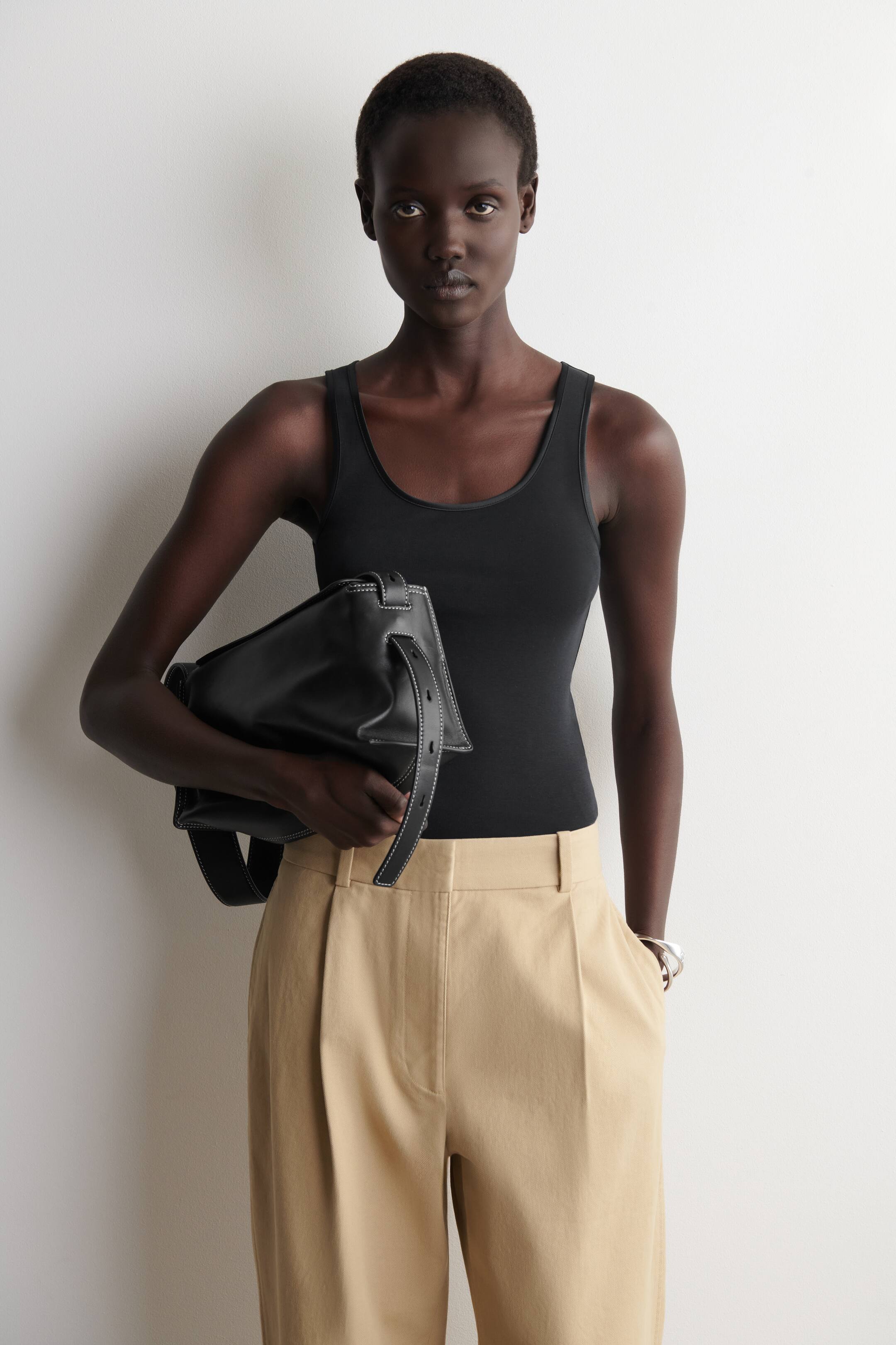 הצגת תמונה גדולה יותר: חולצת טנק שחורה עם מחשוף עגול ומותאם מוצגת כשהיא תחובה בתוך מכנסיים בגזרה גבוהה בצבע בז'. המכנסיים כוללים קפלים בודדים, לולאות לחגורה וגזרה נינוחה.