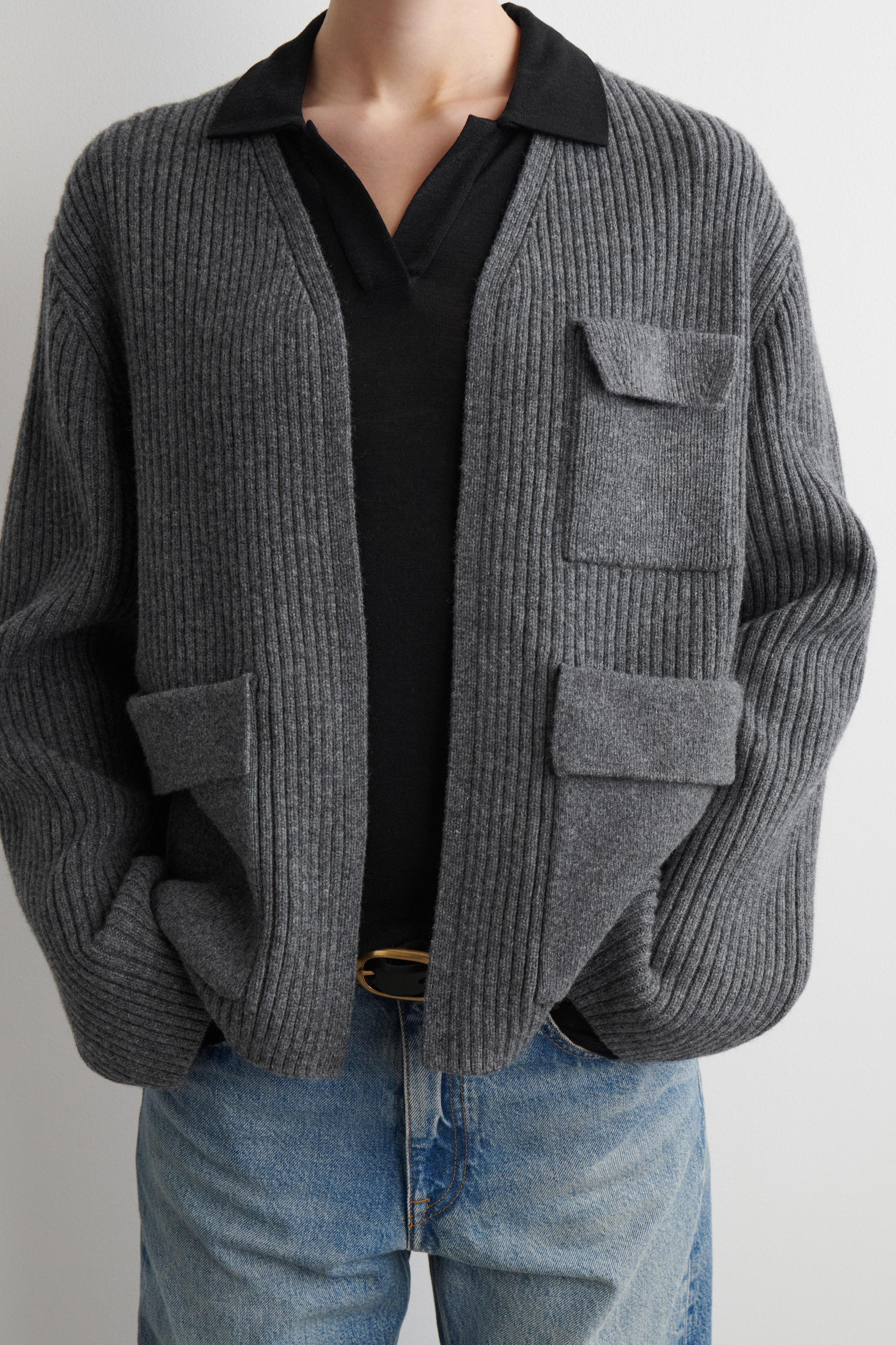 Visualizza immagine più grande: Un cardigan grigio a maglia a coste è indossato aperto, rivolto in avanti. Presenta una vestibilità rilassata, due tasche a toppa sul petto con patta e due tasche a toppa inferiori più grandi con patta, abbinato a un top nero con colletto e jeans azzurri.