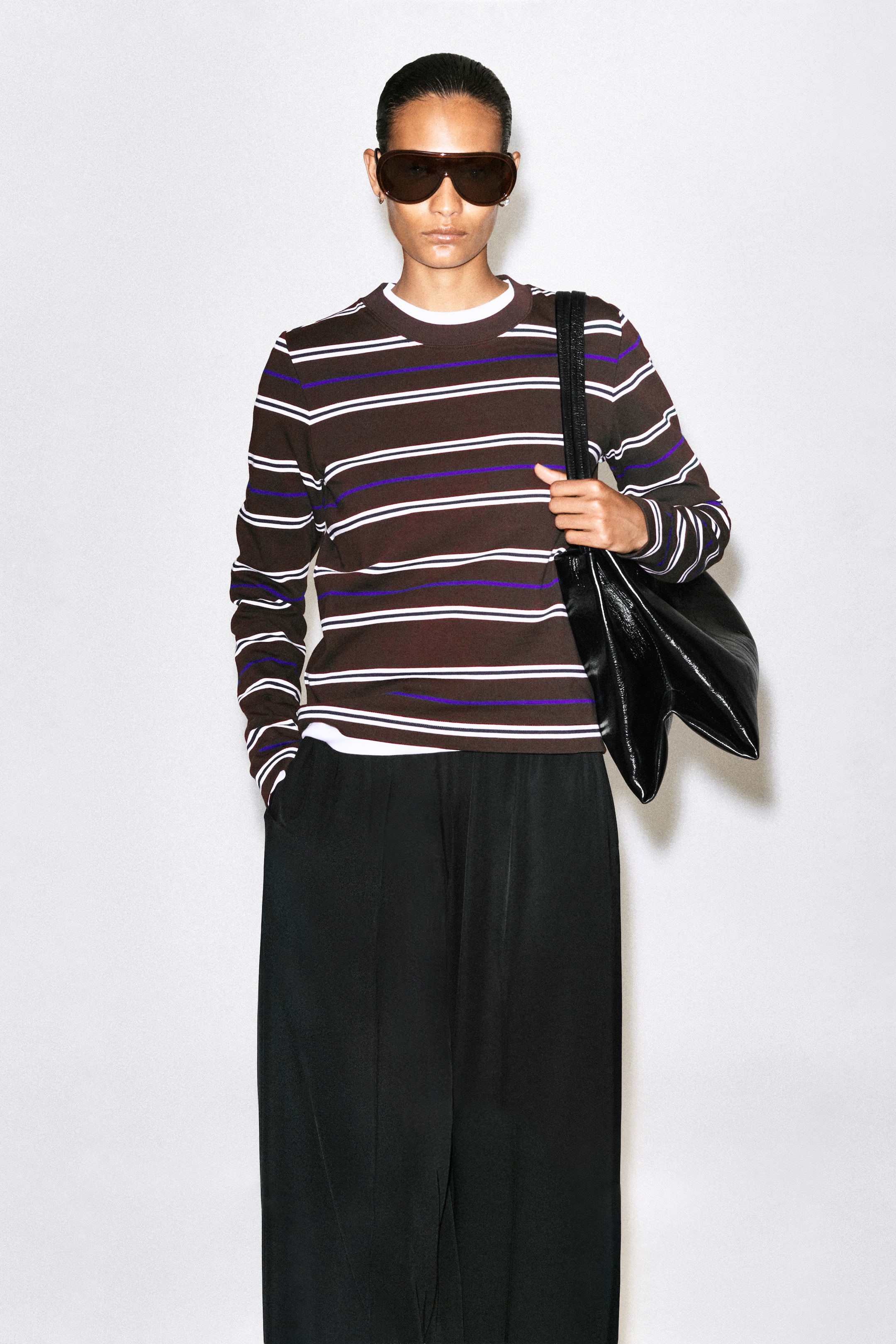 Größeres Bild anzeigen: Ein brauner Pullover mit Rundhalsausschnitt und langen Ärmeln mit horizontalen weißen und violetten Streifen wird vom Model über einem weißen T-Shirt mit Rundhalsausschnitt getragen. Das Oberteil wird mit einer schwarzen Wide-Leg-Hose, dunkler Sonnenbrille und einer schwarzen Umhängetasche kombiniert, die über einer Schulter getragen wird.