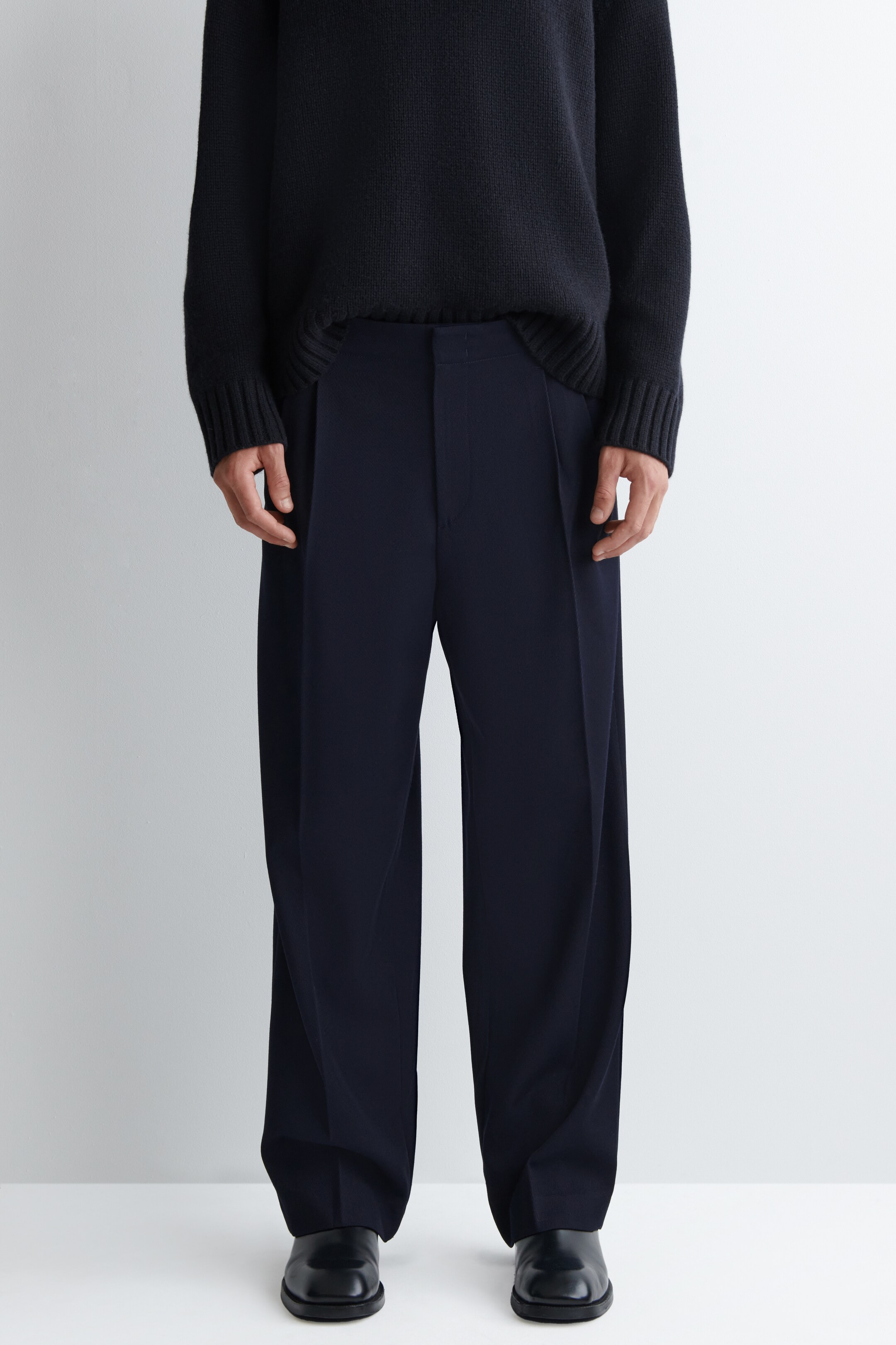 View larger image: Dark blue wide-leg trousers are being shown full-length, facing forward. The trousers are featuring a relaxed fit with single pleats at the high waist and are draping loosely to the floor.