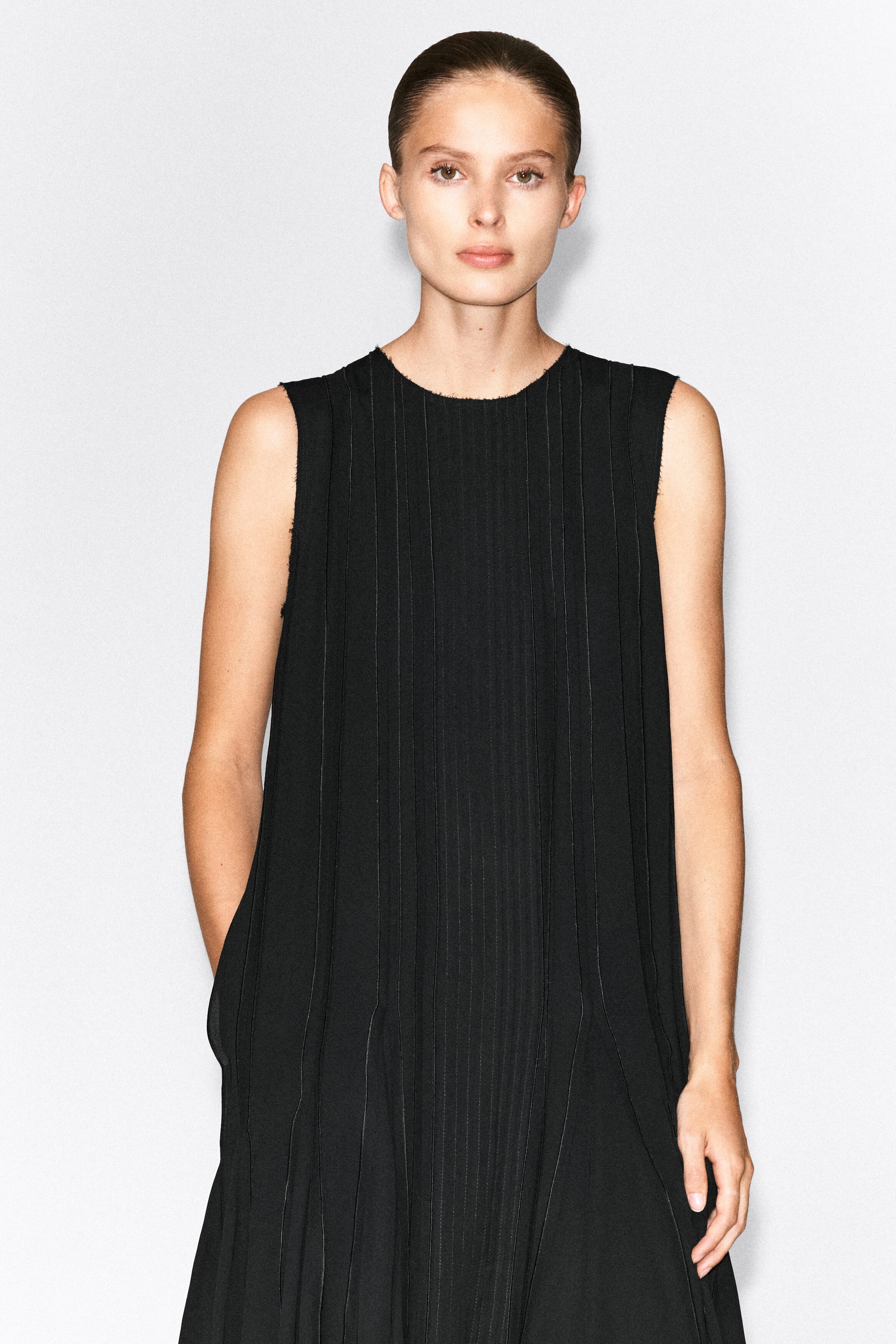 A black, sleeveless midi dress featuring vertical pleated panels and raw-edged trim at the neckline and armholes is being worn by the model, who is resting one hand in a side pocket. A black, sleeveless midi dress featuring vertical pleated panels and raw-edged trim at the neckline and armholes is being worn by the model, who is resting one hand in a side pocket.