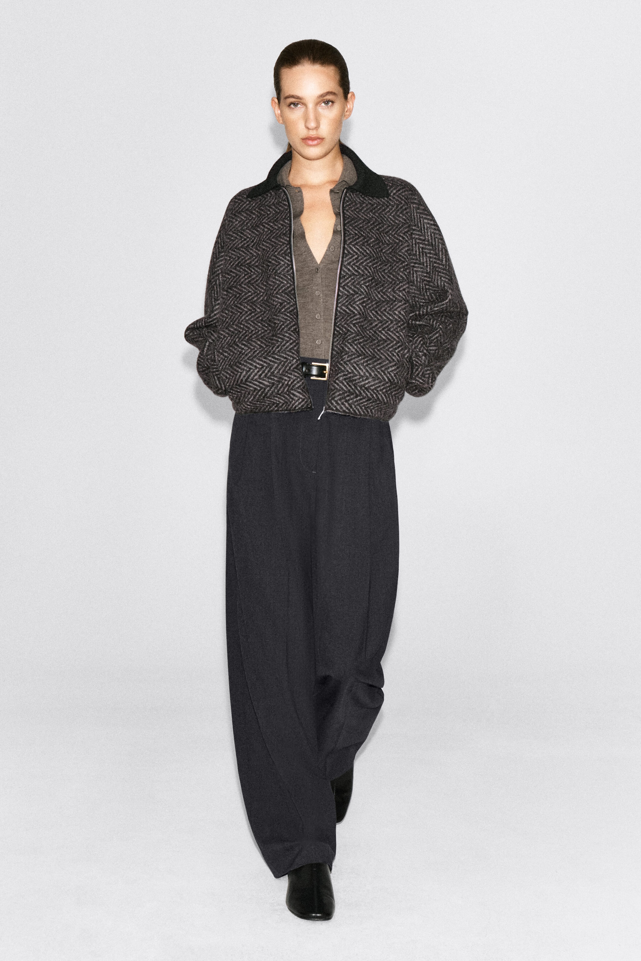Une veste en tricot à motif chevron, dotée d’un col côtelé noir et d’une fermeture à glissière de ton argenté, est portée par le modèle, qui se tient debout, les mains dans les poches latérales. La veste à coupe décontractée est superposée à un haut gris boutonné et à un pantalon foncé à jambes larges.