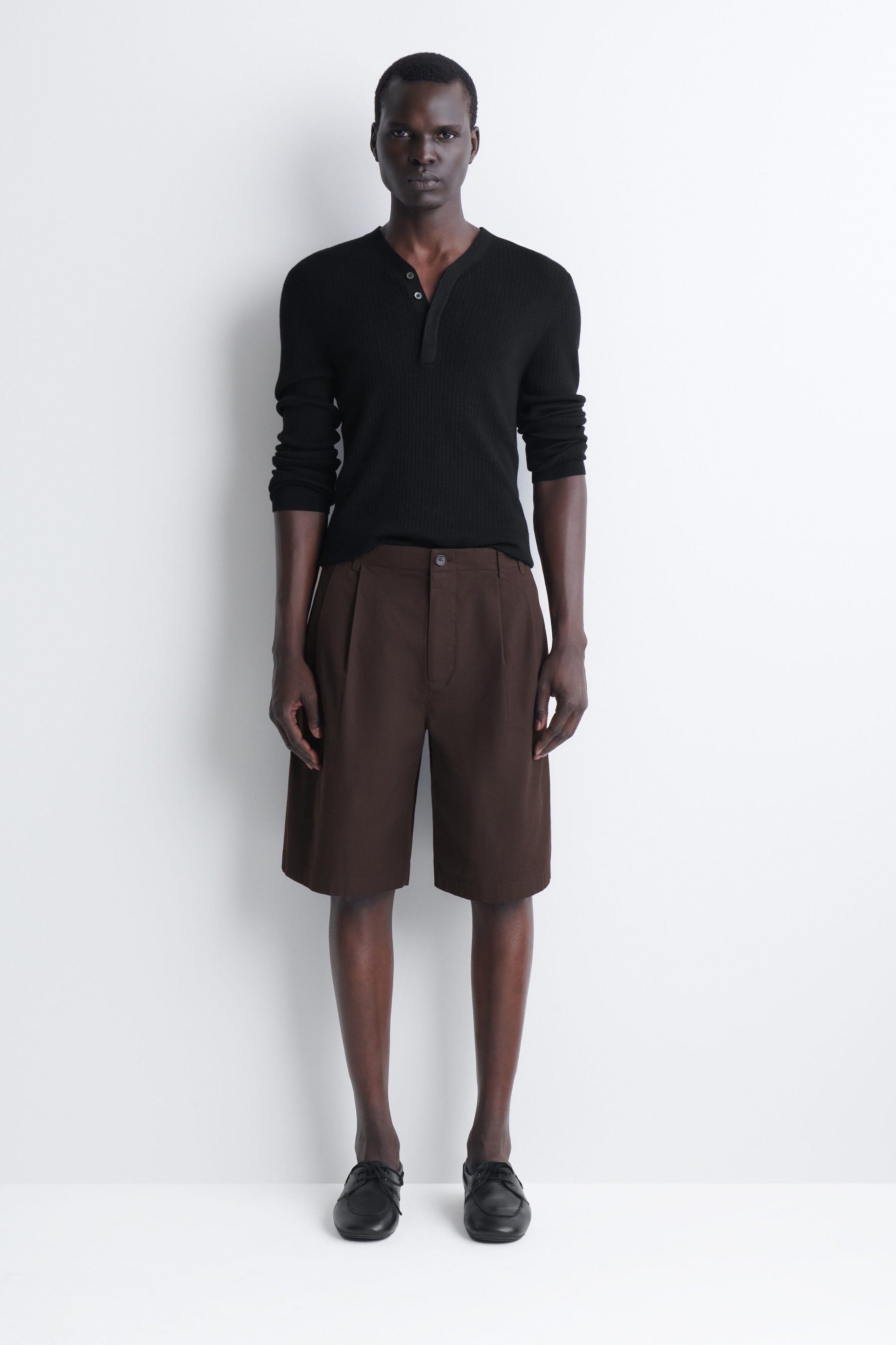 Tailored pleated cotton shorts in dark brown are displayed from the front, featuring a structured waistband with a button fastening and prominent front pleats. The shorts fall to a wide-leg, knee-length fit and are styled with a black Henley shirt tucked in to emphasise the waist. Tailored pleated cotton shorts in dark brown are displayed from the front, featuring a structured waistband with a button fastening and prominent front pleats. The shorts fall to a wide-leg, knee-length fit and are styled with a black Henley shirt tucked in to emphasise the waist.