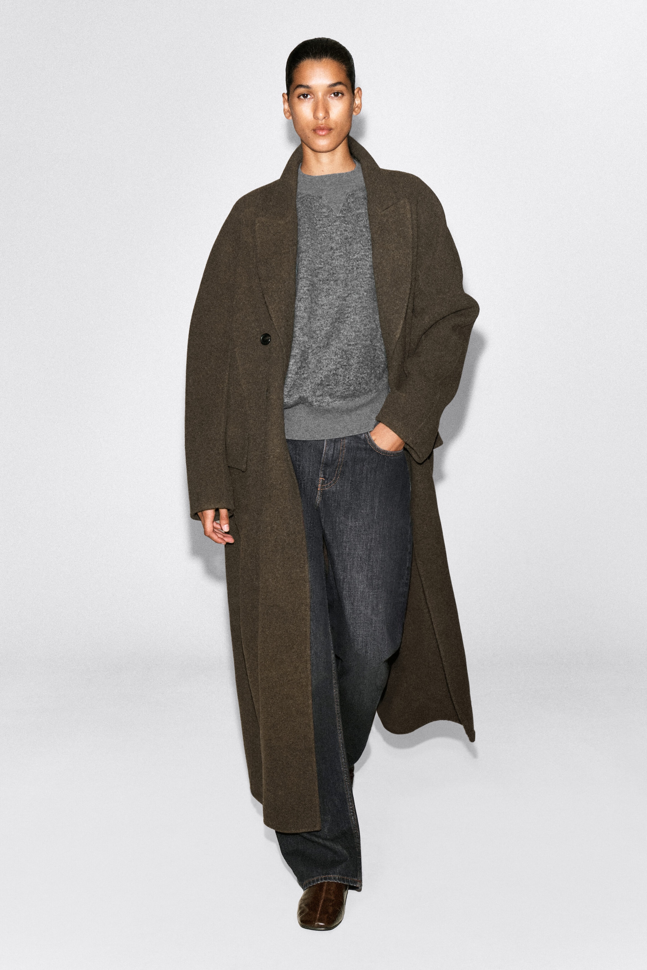 A long, oversized wool coat in a dark olive-brown color is shown from the front, featuring wide notch lapels, dropped shoulders, and a relaxed, ankle-length silhouette. The coat is styled open over a gray textured sweater and dark jeans to highlight its fluid drape and roomy fit. A long, oversized wool coat in a dark olive-brown color is shown from the front, featuring wide notch lapels, dropped shoulders, and a relaxed, ankle-length silhouette. The coat is styled open over a gray textured sweater and dark jeans to highlight its fluid drape and roomy fit.