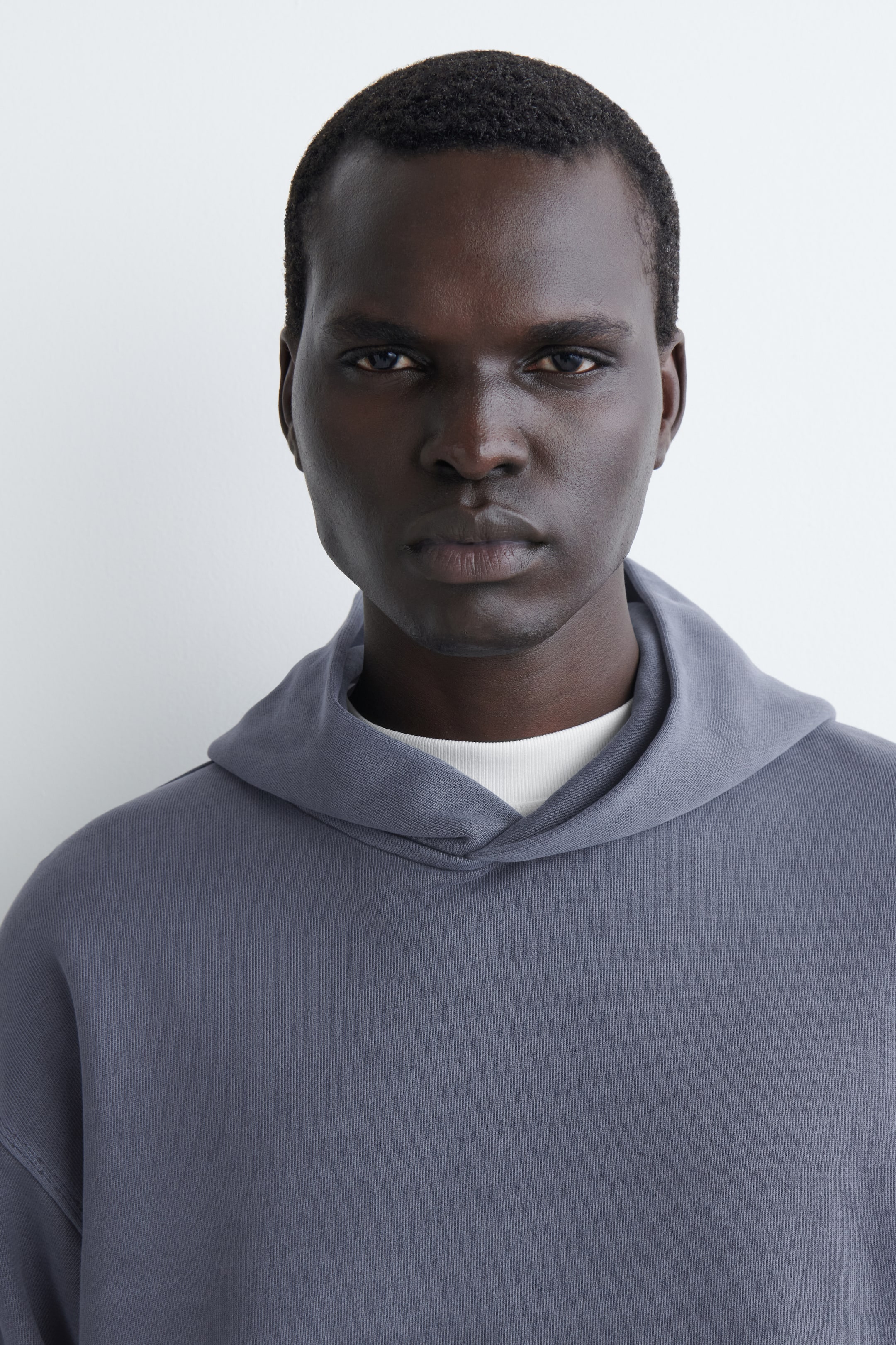 Grösseres Bild ansehen: Ein schiefergrauer Hoodie wird von vorne gezeigt. Er besteht aus einem weichen, strukturierten Stoff, einer drapierten Kapuze und einem darunter sichtbaren weißen Rundhals-Shirt.