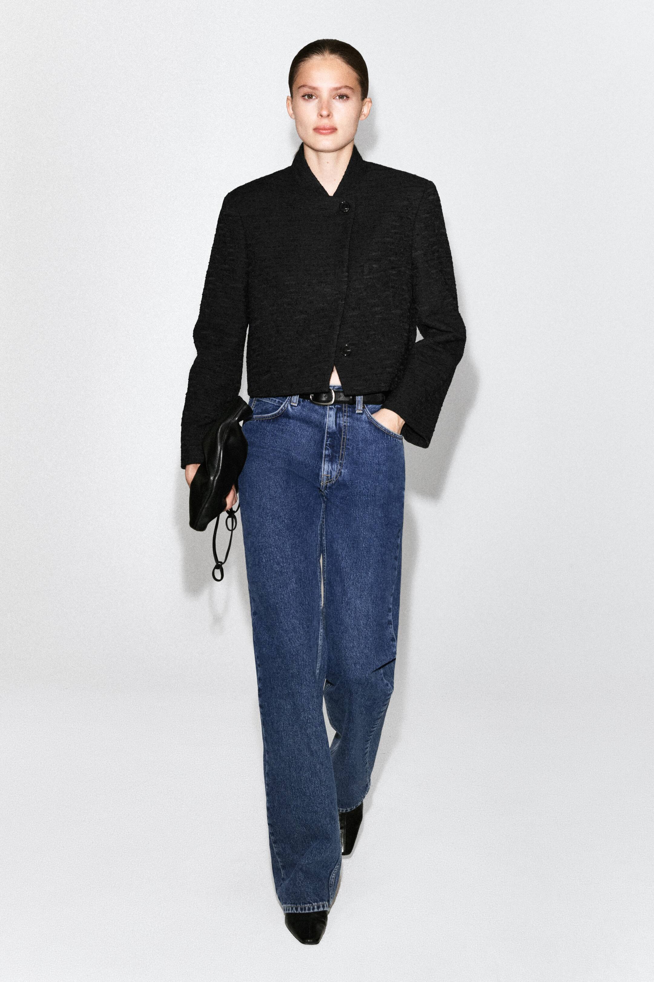 View larger image: A black textured cropped jacket with a Mandarin collar and front button fastening is being worn by the model. It is styled with blue jeans and a black belt.