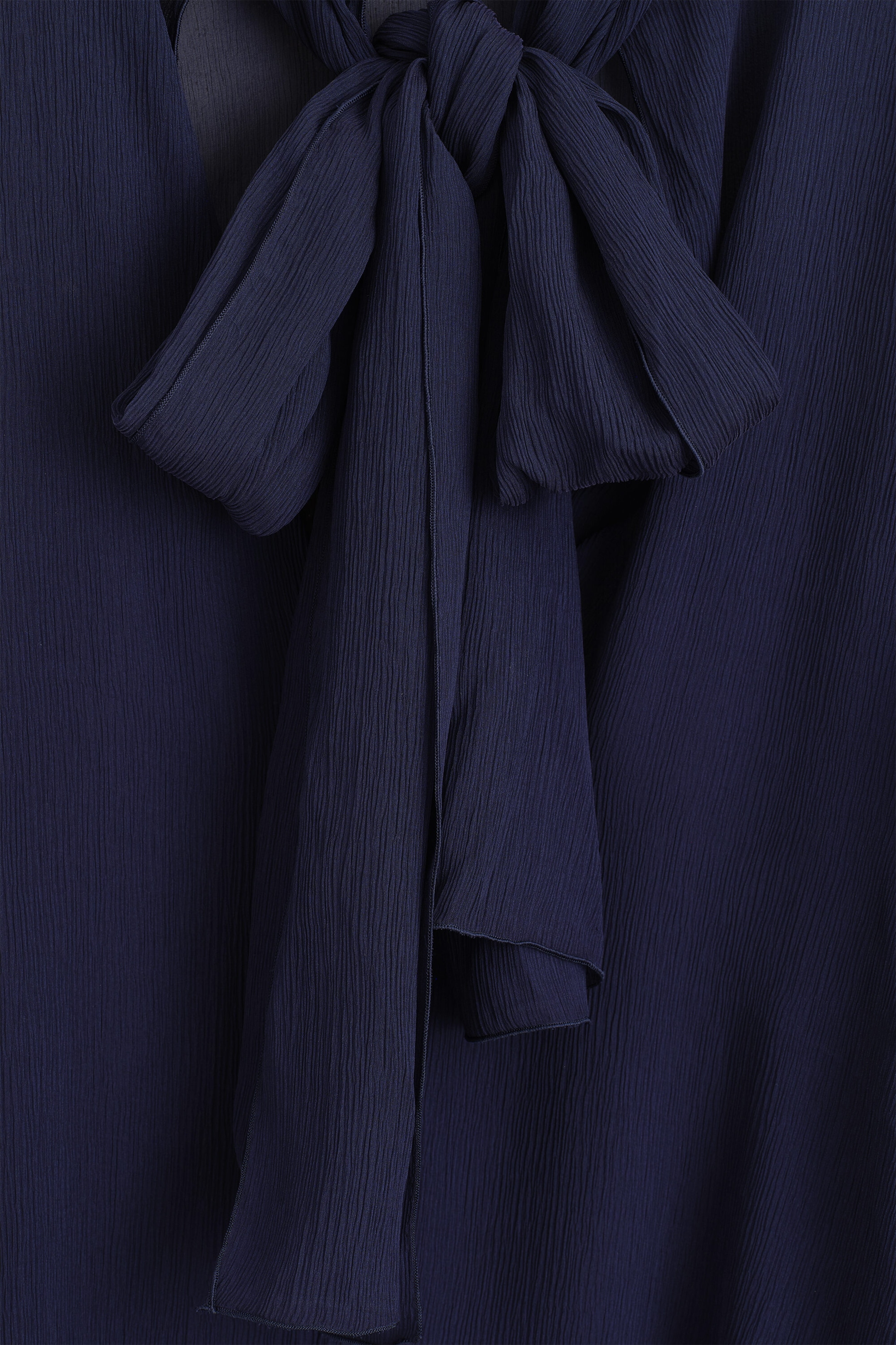 A close-up front view showing a navy blue crepe top featuring a self-tie scarf detail at the neckline. The scarf is draped in a loose knot, showcasing the fine crinkled texture of the semi-sheer fabric. A close-up front view showing a navy blue crepe top featuring a self-tie scarf detail at the neckline. The scarf is draped in a loose knot, showcasing the fine crinkled texture of the semi-sheer fabric.