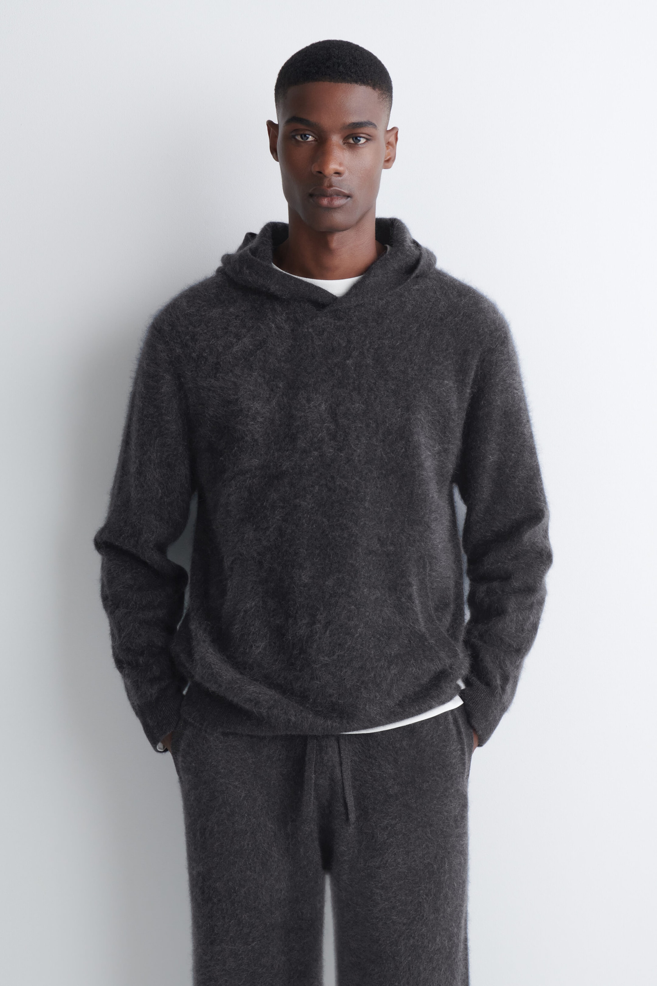 The model is wearing a dark gray brushed cashmere hoodie with a soft, textured finish. The hoodie is layered over a white T-shirt and paired with matching dark gray knit trousers.