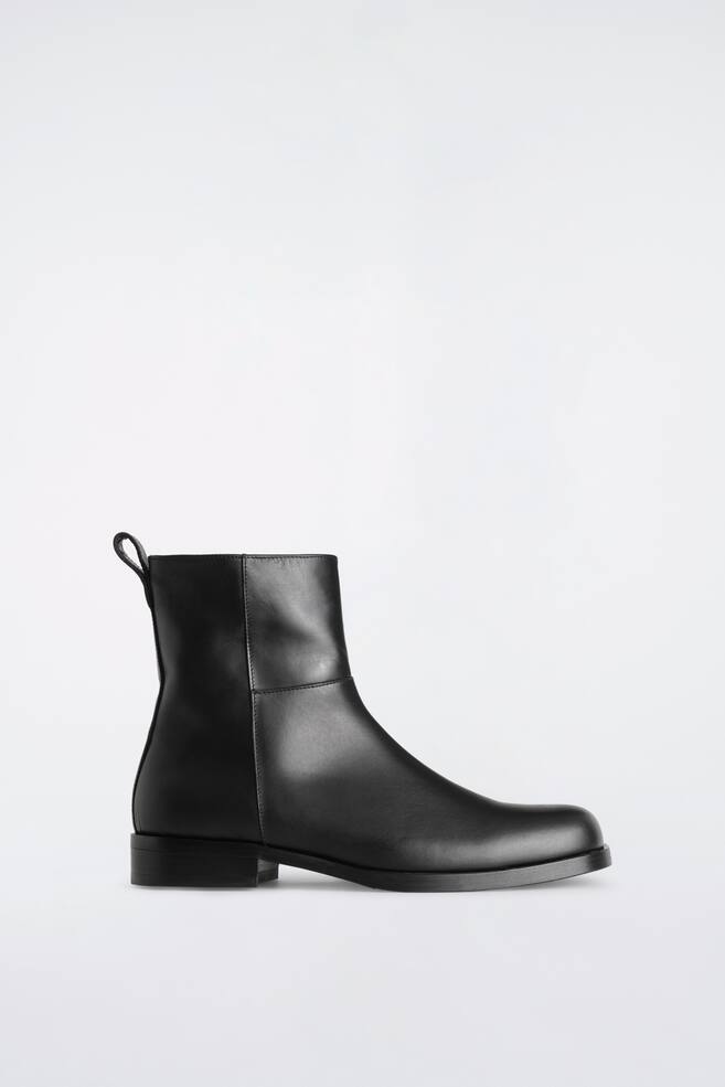 Ein schwarzer Leder-Ankle-Boot ist in der Seitenansicht zu sehen und prÃ¤sentiert ein glattes, mattes Finish, eine abgerundete Zehenpartie und einen niedrigen Blockabsatz. Der Boot zeichnet sich durch ein minimalistisches Design mit sauberen tonalen NÃ¤hten und einer Zuglasche am hinteren Schaftabschluss aus. - 1