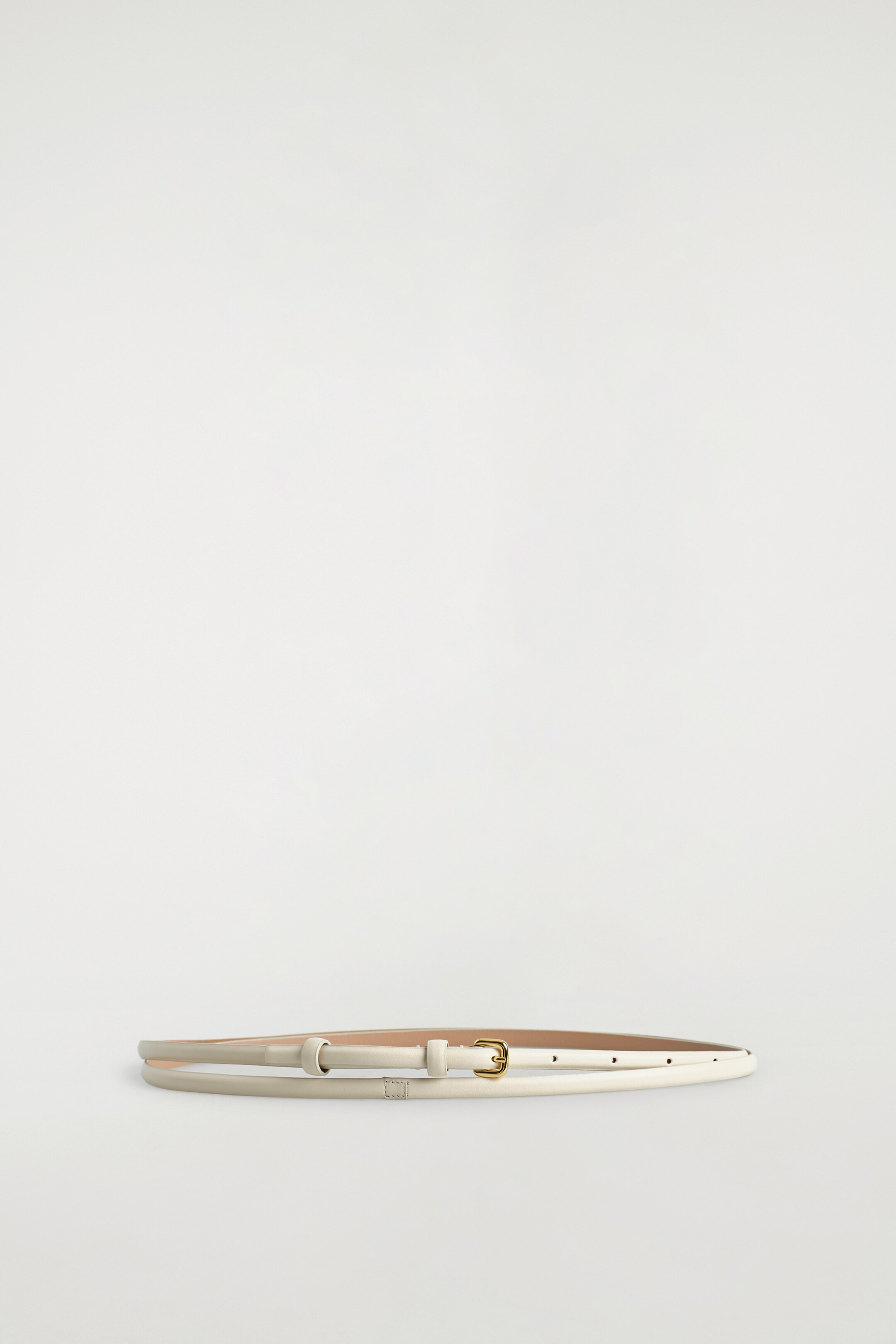 Zobrazit větší obrázek: Úzký, světle béžový pásek leží naplocho a je stočený do oválného tvaru. Má malou, zlatě tónovanou, organicky tvarovanou přezku a jednu poutko na pásek.