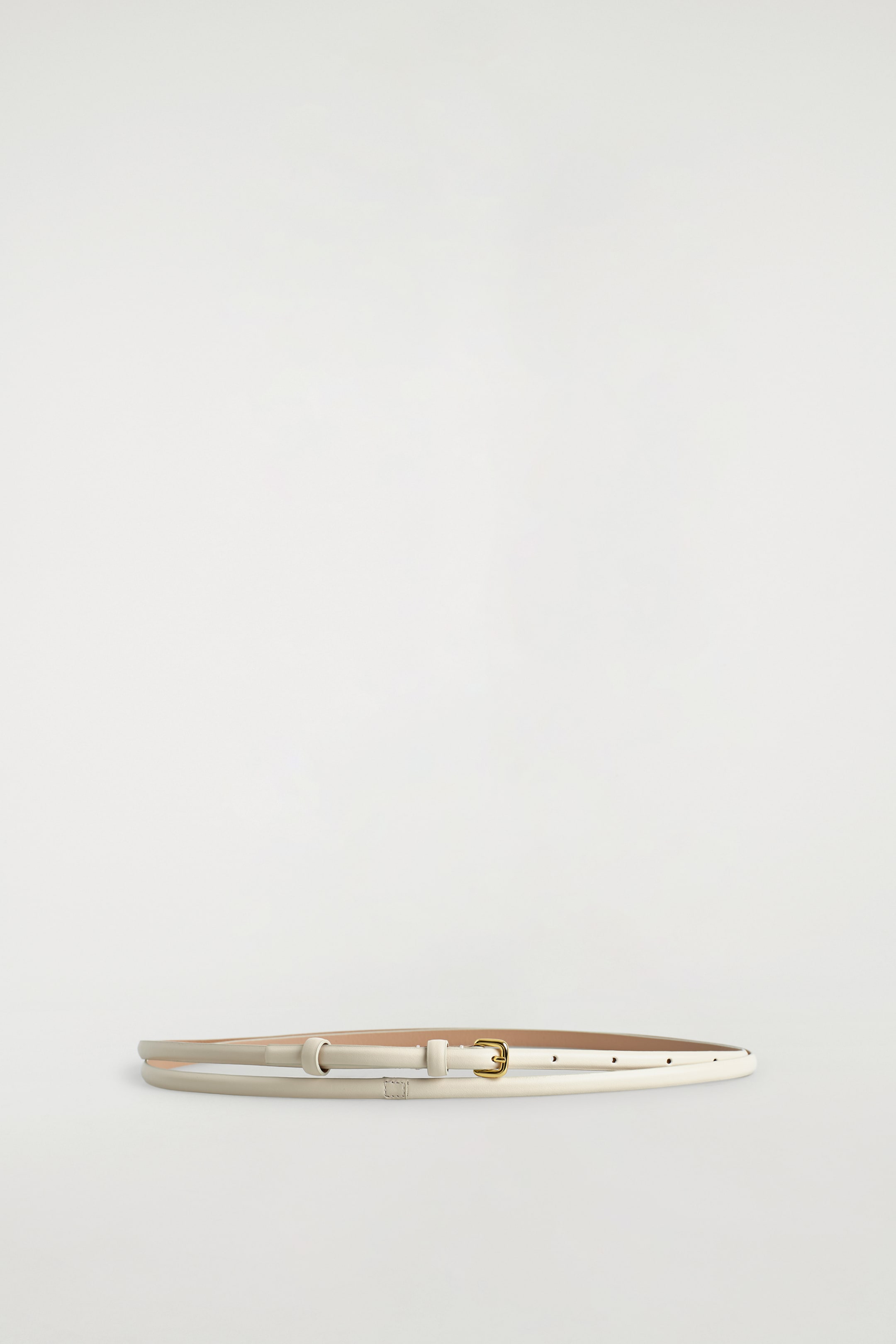 Agrandir l'image: Une ceinture fine, de couleur beige clair, est posée à plat et enroulée en forme ovale. Elle est dotée d'une petite boucle dorée de forme organique et d'un seul passant de ceinture.