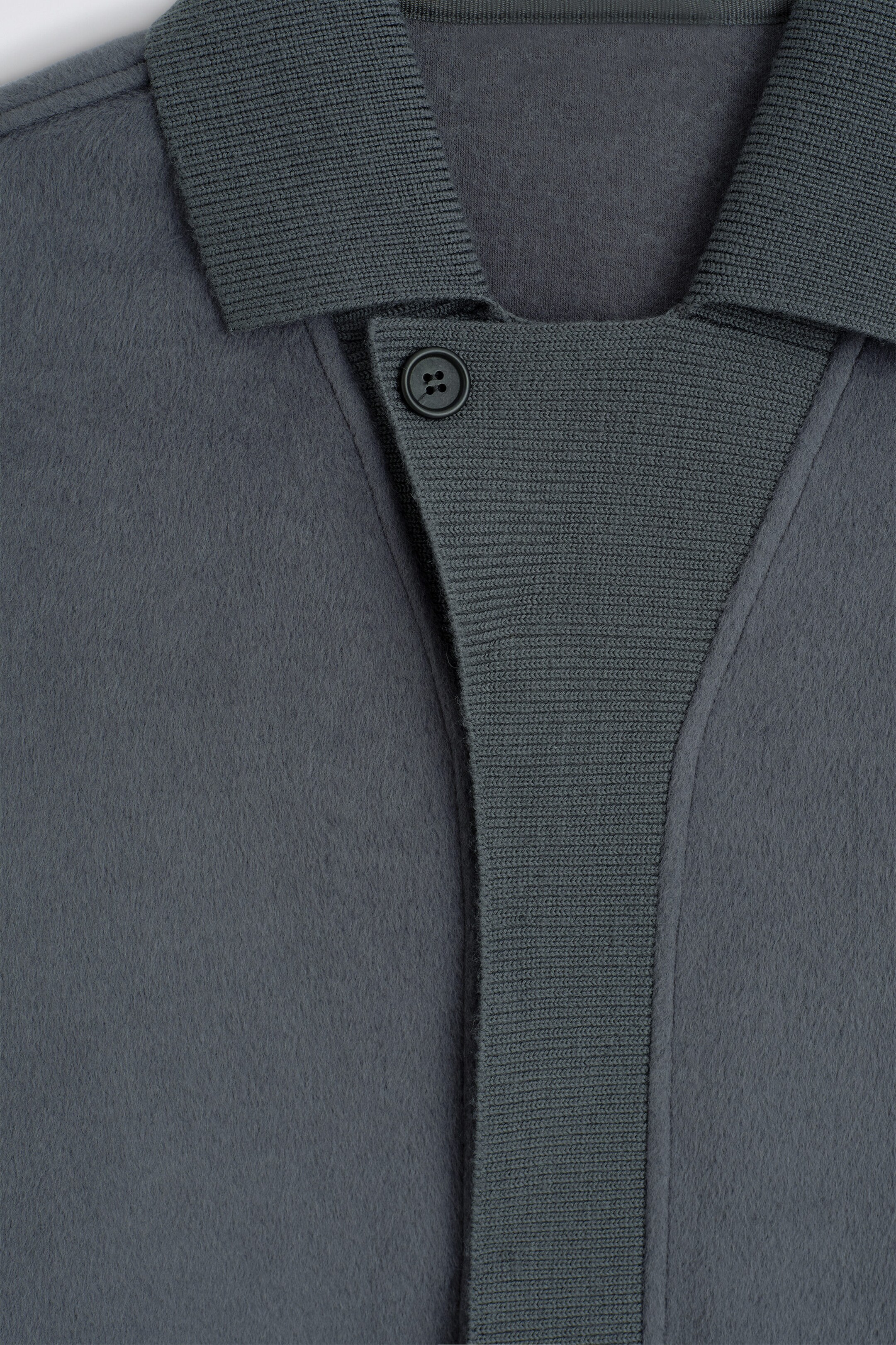 Une chemise en tricot gris foncé présente une texture douce et brossée sur le corps, contrastée par un col en tricot côtelé et une patte de boutonnage avant côtelée assortie. Un seul bouton foncé ferme le haut de la patte de boutonnage dans cette vue rapprochée de face. Une chemise en tricot gris foncé présente une texture douce et brossée sur le corps, contrastée par un col en tricot côtelé et une patte de boutonnage avant côtelée assortie. Un seul bouton foncé ferme le haut de la patte de boutonnage dans cette vue rapprochée de face.