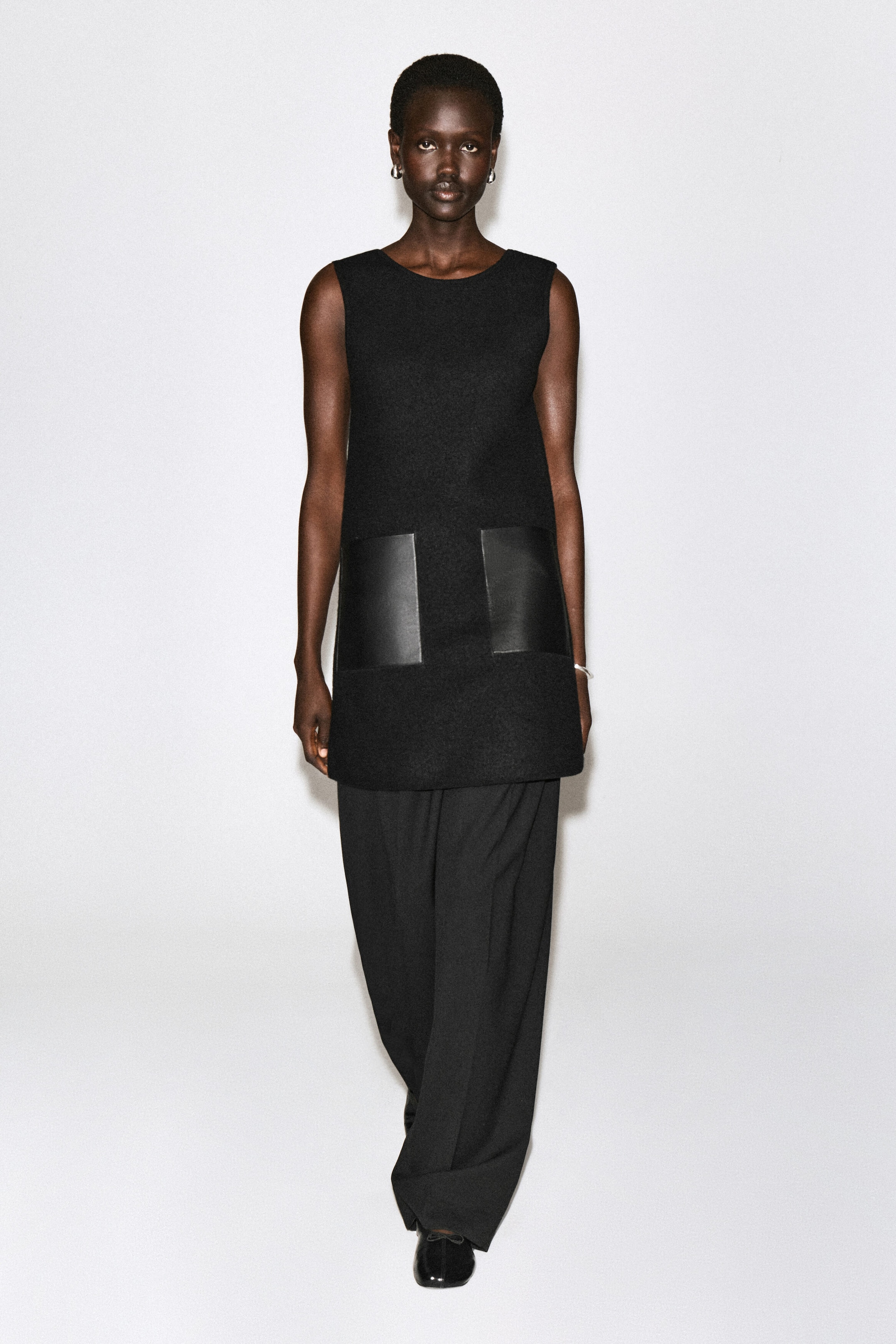 A black sleeveless mini dress is shown from the front, featuring a structured shift silhouette, a crew neckline, and two large square leather patch pockets on the lower front. The dress is styled as a tunic layered over wide-leg black trousers. A black sleeveless mini dress is shown from the front, featuring a structured shift silhouette, a crew neckline, and two large square leather patch pockets on the lower front. The dress is styled as a tunic layered over wide-leg black trousers.