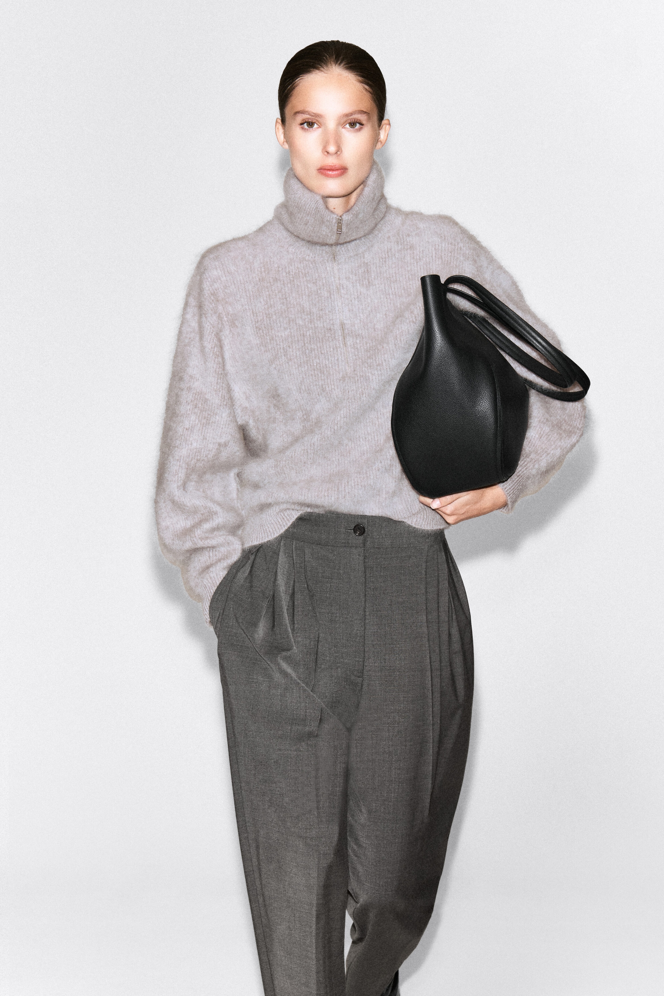 A light gray brushed cashmere sweater with a high funnel neck and a silver-tone zipper is worn by the model. The sweater is partially tucked into dark gray pleated trousers, with a black leather handbag tucked under one arm. A light gray brushed cashmere sweater with a high funnel neck and a silver-tone zipper is worn by the model. The sweater is partially tucked into dark gray pleated trousers, with a black leather handbag tucked under one arm.
