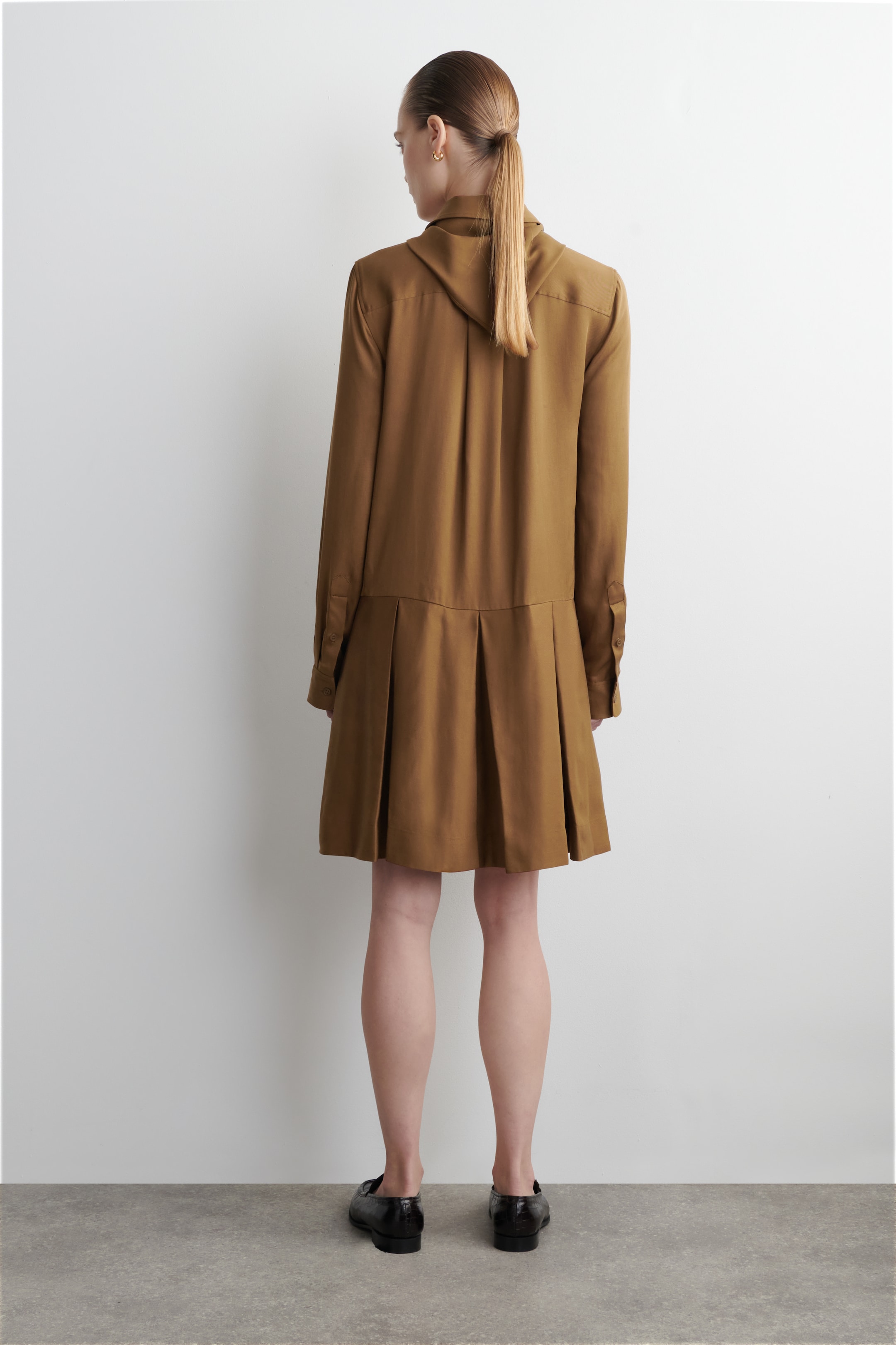 Ver imagem maior: Um vestido tipo camisa de cor camel está a ser apresentado de costas, com um detalhe de decote drapeado e mangas compridas com punhos abotoados. A parte superior das costas exibe pregas verticais, e uma saia plissada estende-se a partir de uma costura horizontal na cintura.