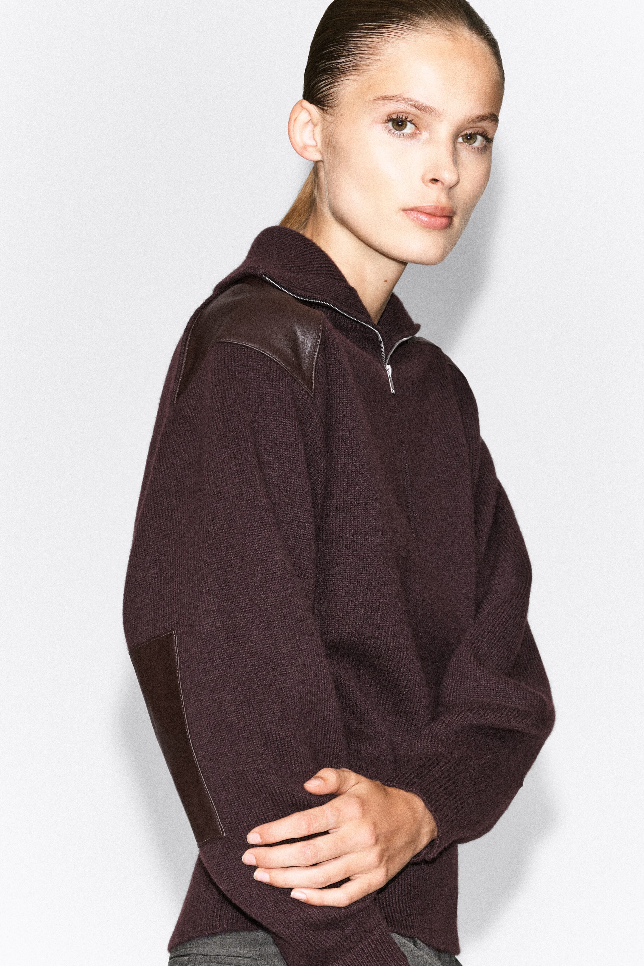 The model is wearing a dark burgundy merino wool zip-up sweater, with one elbow bent and held in place. The sweater features a silver half-zip collar, a brown leather patch on the shoulder, and a matching leather patch on the elbow. The model is wearing a dark burgundy merino wool zip-up sweater, with one elbow bent and held in place. The sweater features a silver half-zip collar, a brown leather patch on the shoulder, and a matching leather patch on the elbow.