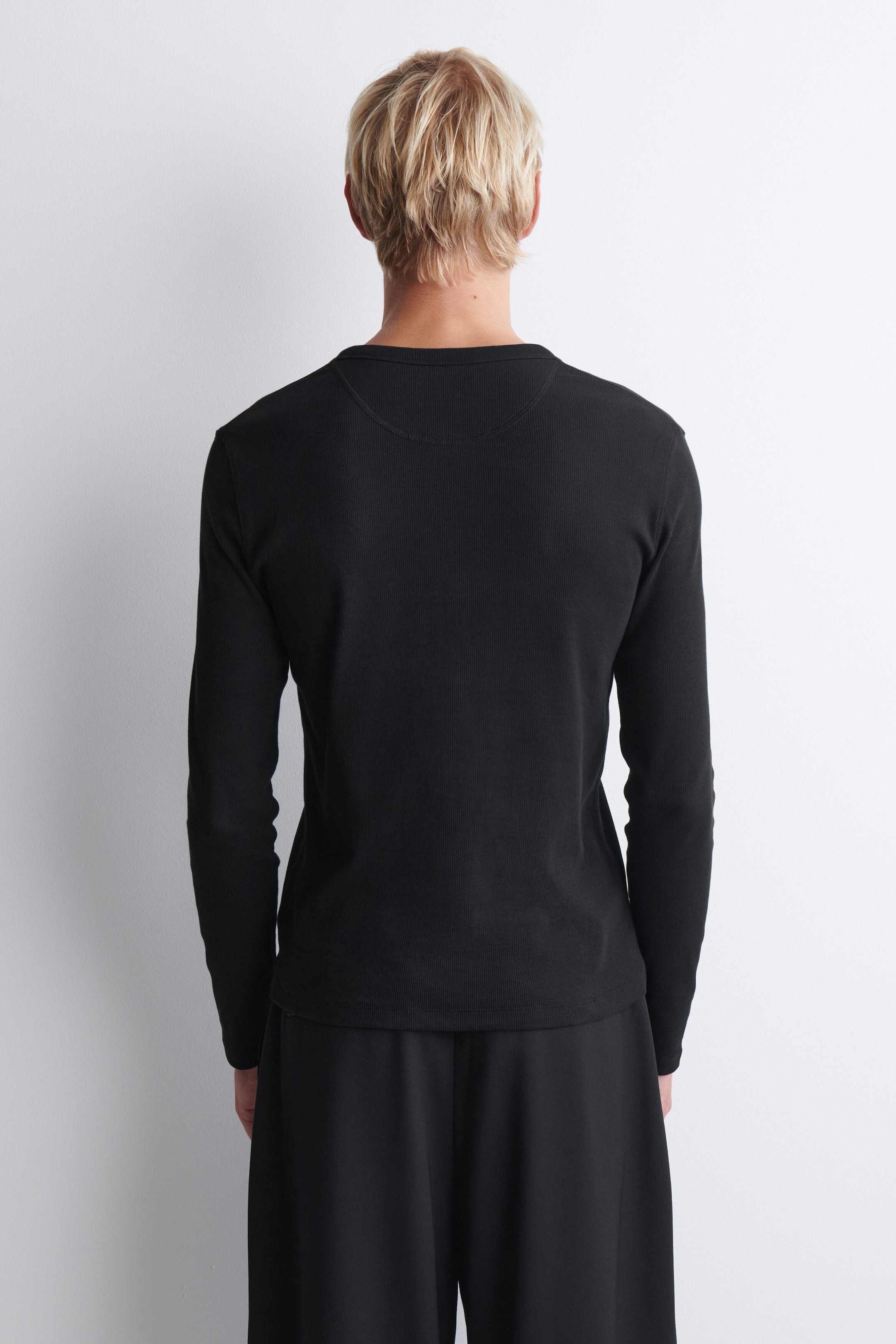 The slim ribbed cotton long-sleeve Henley T-shirt is shown from the back, highlighting its close-fitting design, straight hem, and curved yoke seam below the crew neckline. The black long-sleeve shirt is paired with matching black trousers to showcase its fit and length. The slim ribbed cotton long-sleeve Henley T-shirt is shown from the back, highlighting its close-fitting design, straight hem, and curved yoke seam below the crew neckline. The black long-sleeve shirt is paired with matching black trousers to showcase its fit and length.
