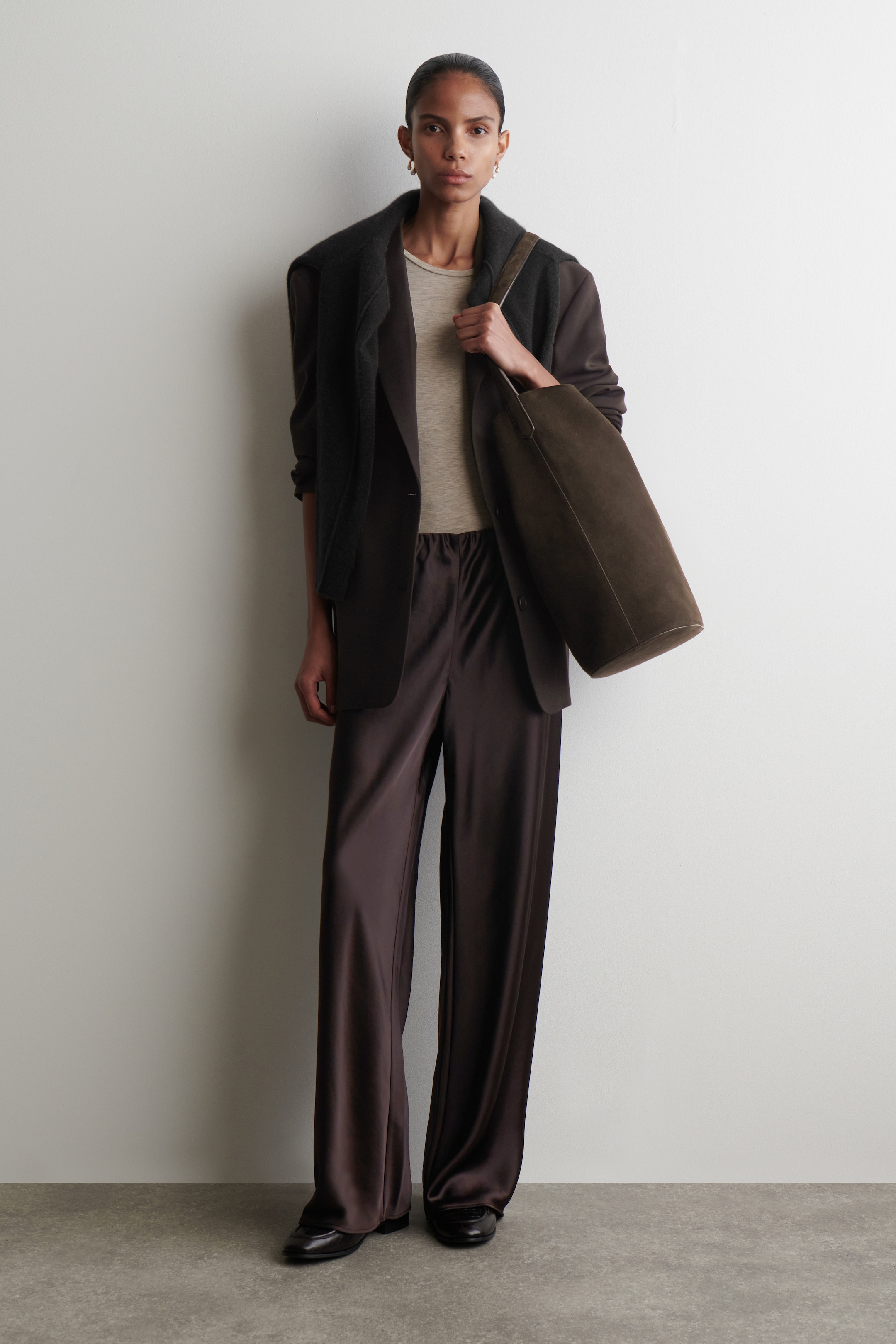 Visualizza immagine più grande: Il prodotto è un completo che presenta un top girocollo beige chiaro, un blazer monopetto marrone scuro e pantaloni marrone scuro a gamba larga in satin con elastico in vita, il tutto con una vestibilità rilassata. Un maglione in maglia color carbone è drappeggiato sulle spalle, una borsa tote di camoscio verde oliva scuro viene tenuta in mano e scarpe piatte nere sono indossate ai piedi.