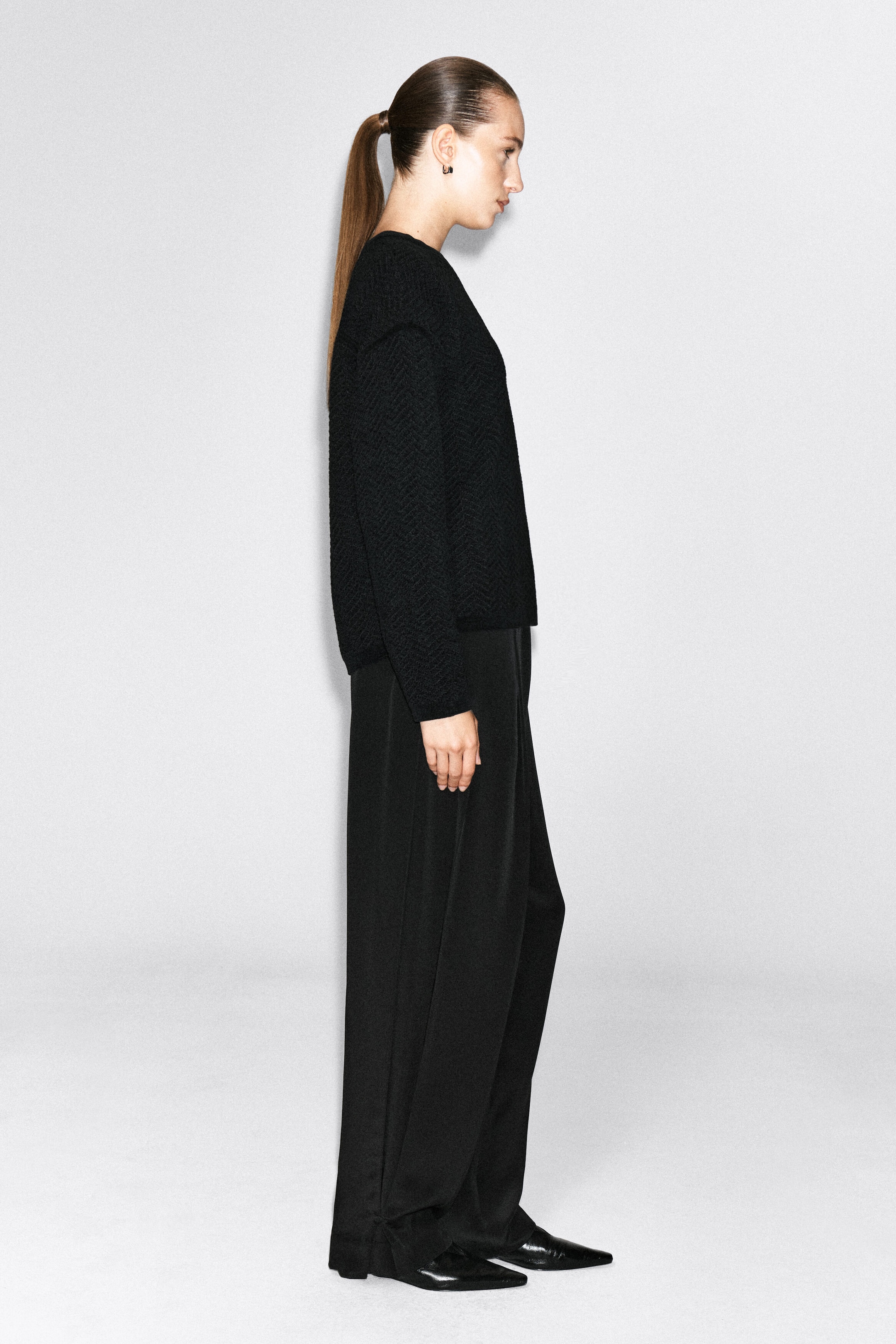 This black merino wool chenille crew-neck sweater is worn by the model, highlighting its textured knit pattern, long sleeves, and relaxed silhouette. It is styled with matching black wide-leg trousers and black pointed-toe shoes. This black merino wool chenille crew-neck sweater is worn by the model, highlighting its textured knit pattern, long sleeves, and relaxed silhouette. It is styled with matching black wide-leg trousers and black pointed-toe shoes.