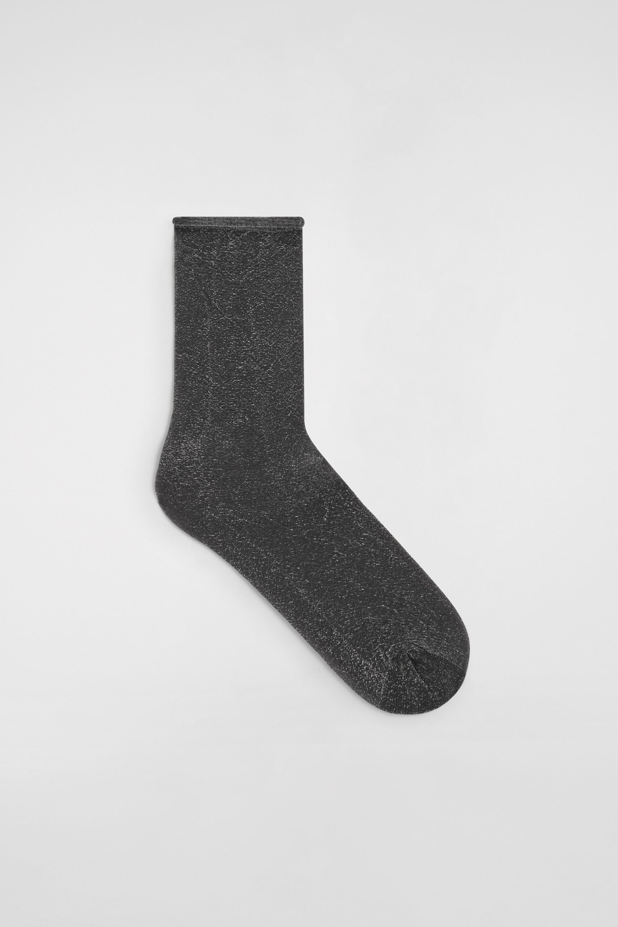 Une chaussette scintillante est posée à plat en vue de profil, mettant en valeur une maille métallique gris foncé parcourue de fils argentés étincelants. La chaussette présente un talon et une pointe structurés, ainsi qu’une fine bordure en haut à bord roulé.
