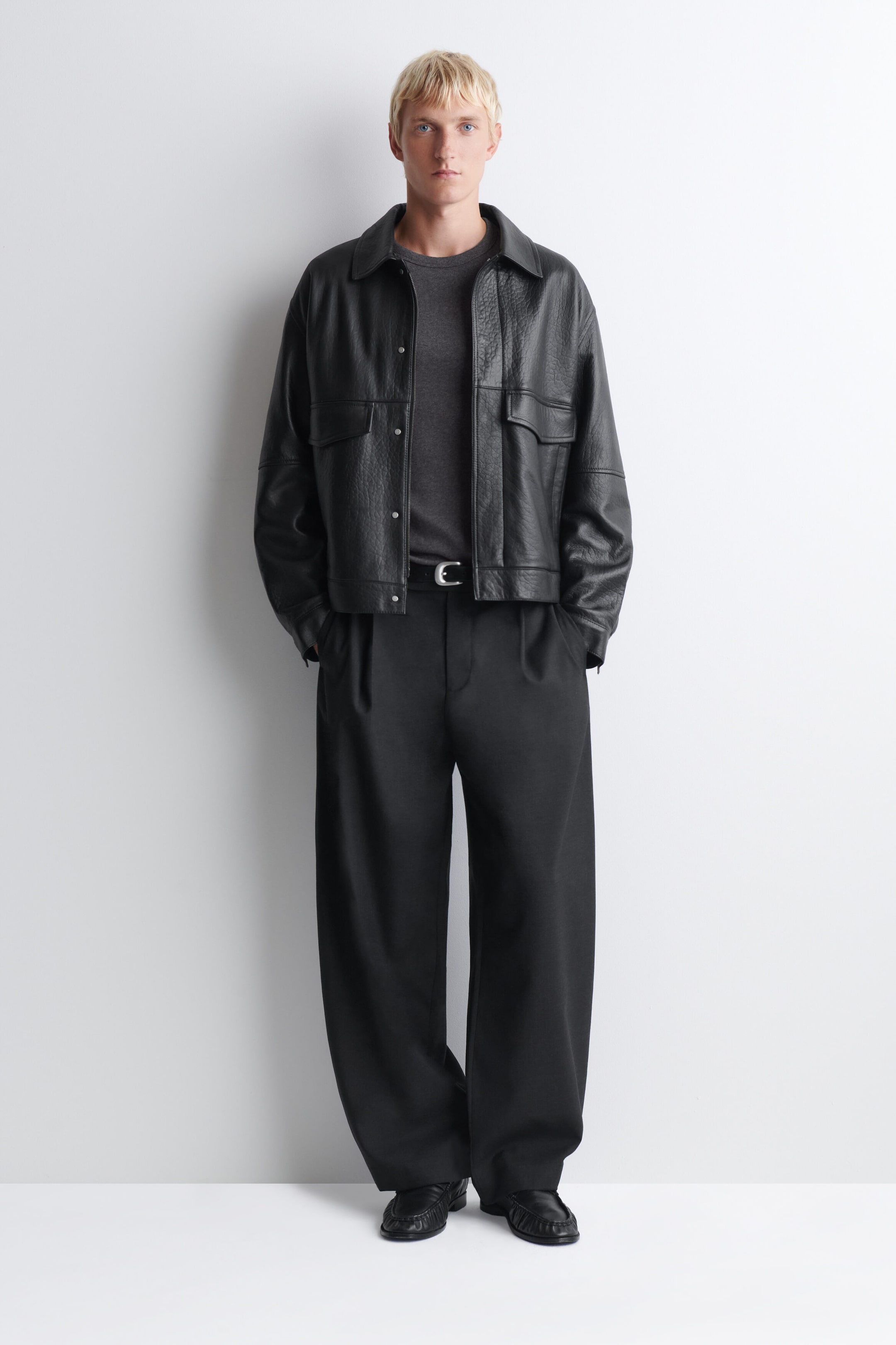 Facing forward, the model is wearing a textured black leather jacket with a pointed collar, snap-button closures, and oversized chest flap pockets over a dark gray crewneck T-shirt. The jacket is styled with black wide-leg pleated trousers, finished with a black belt and a silver-tone buckle.
